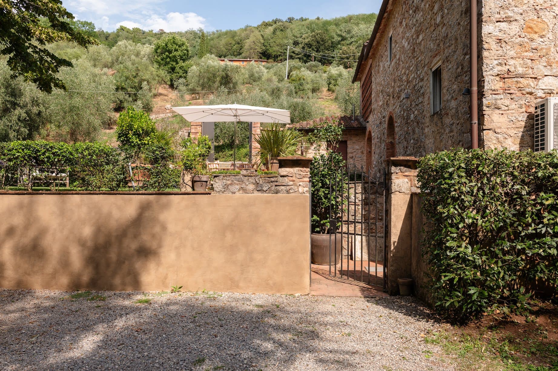 Un bâtiment rustique en pierre se dresse à côté d'une entrée fermée entourée d'une végétation luxuriante. Un haut mur beige et un portail en fer mènent à un patio bien entretenu, doté d'un parasol blanc et de sièges extérieurs. Des collines et des arbres vallonnés remplissent l'arrière-plan sous un ciel partiellement nuageux.