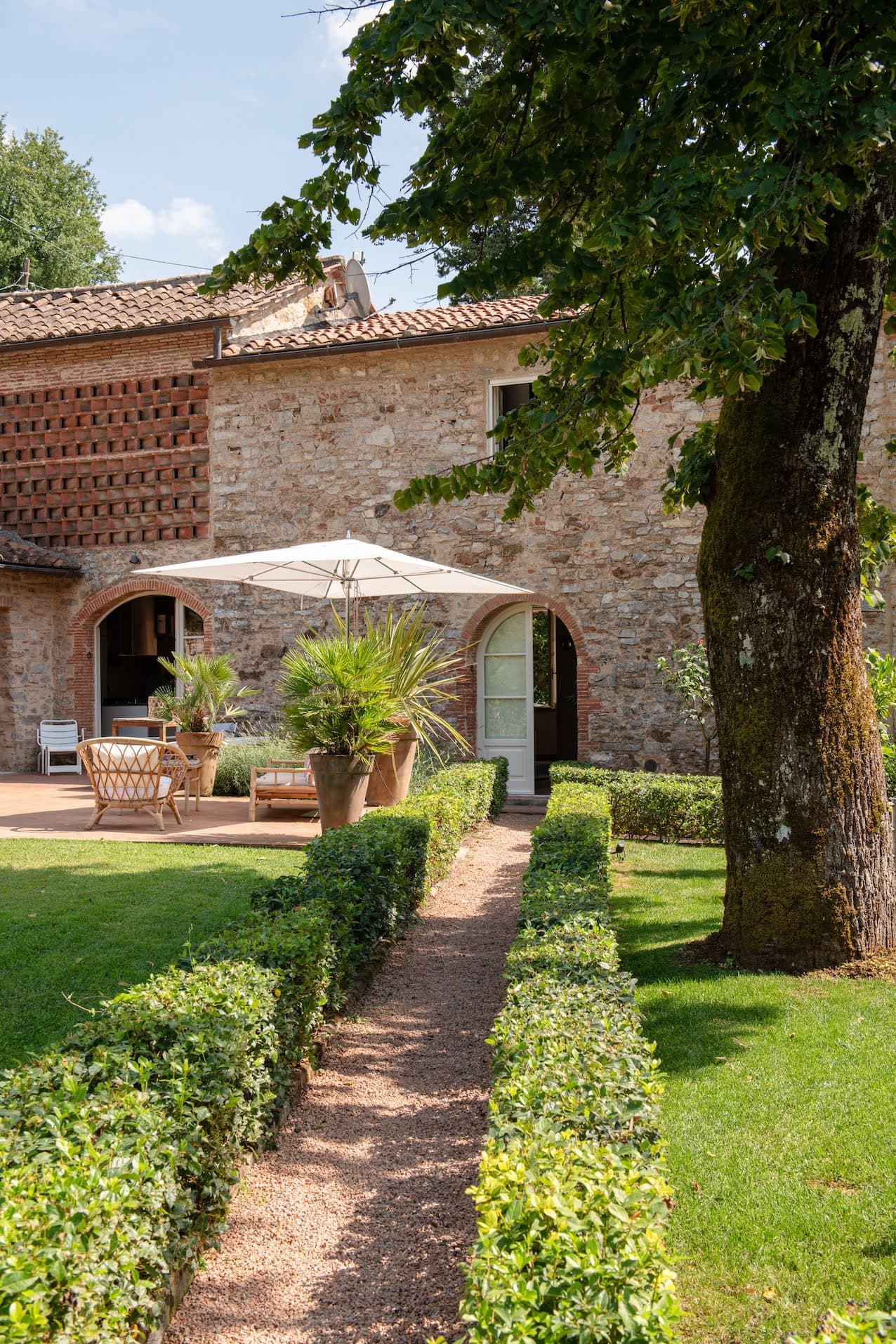Une charmante maison en pierre avec un toit en tuiles de terre cuite se dresse au milieu d'un jardin bien entretenu. Une allée de gravier bordée de haies soigneusement taillées mène à une terrasse avec des meubles en bois et un parasol blanc. De grandes plantes en pot et un arbre rehaussent le cadre serein.