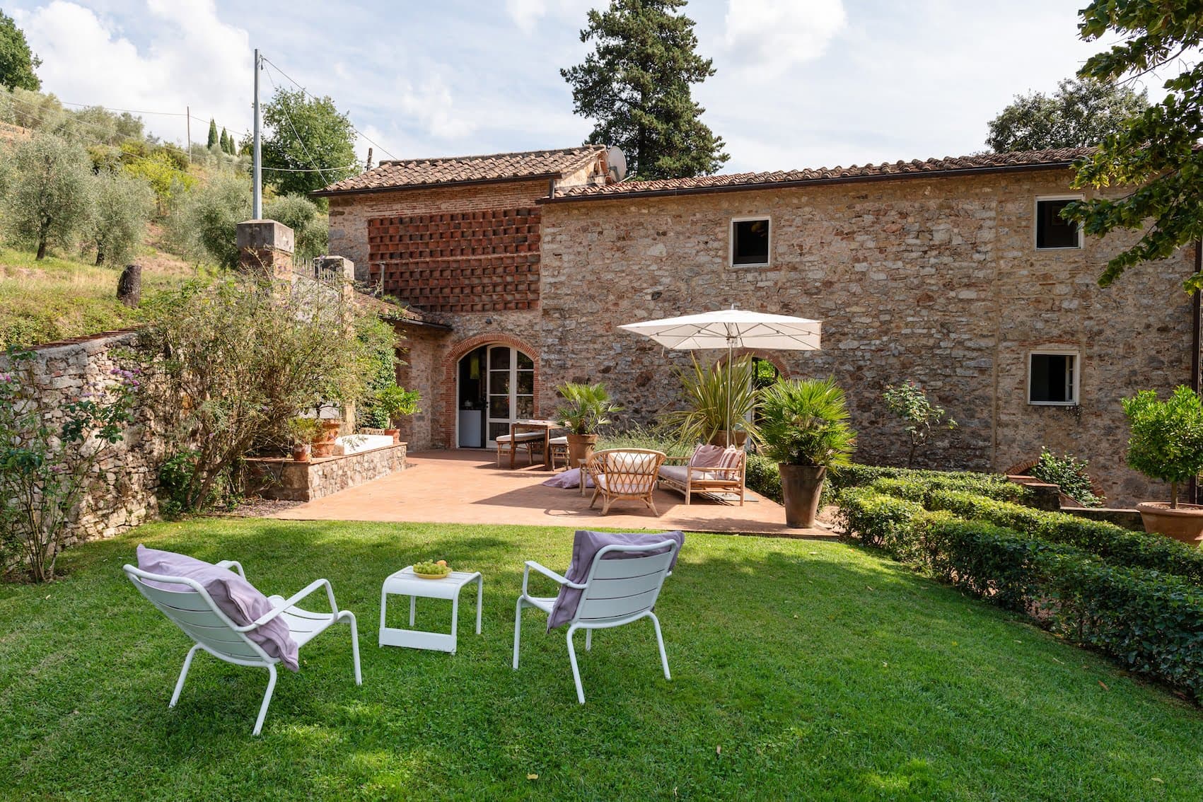 Un charmant patio avec une maison en pierre et un toit en tuiles en arrière-plan. Le patio est doté de meubles d'extérieur, d'un parasol et de plantes en pot. Au premier plan, deux chaises blanches avec coussins et une petite table sont installées sur une pelouse verte et luxuriante.