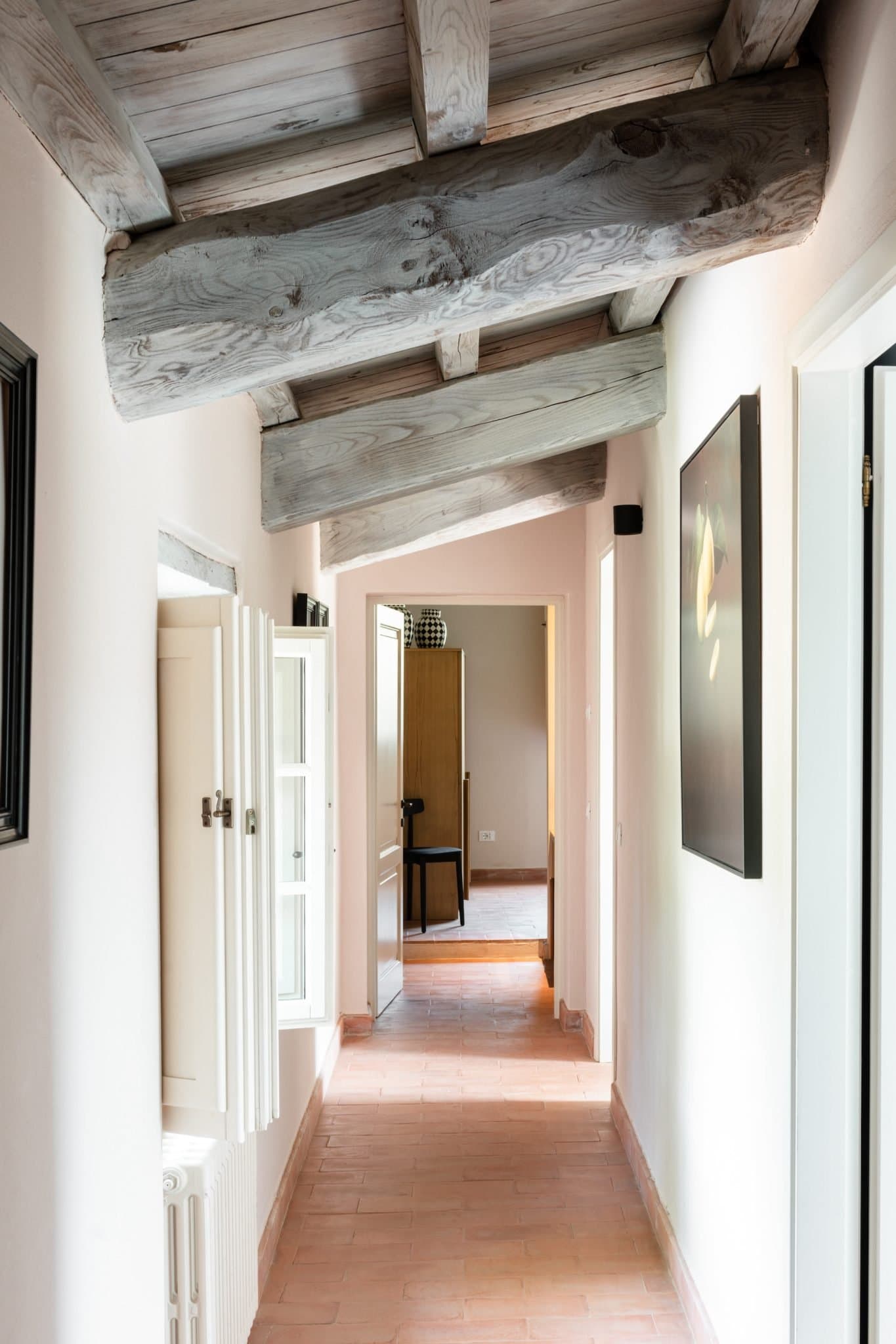 Un couloir avec un plafond à poutres apparentes et des murs de couleur claire. Le sol est carrelé en terre cuite. Sur le mur de gauche, il y a une petite fenêtre et un radiateur blanc. Le mur de droite est orné d'œuvres d'art encadrées. Au bout, une porte ouverte révèle une autre pièce.
