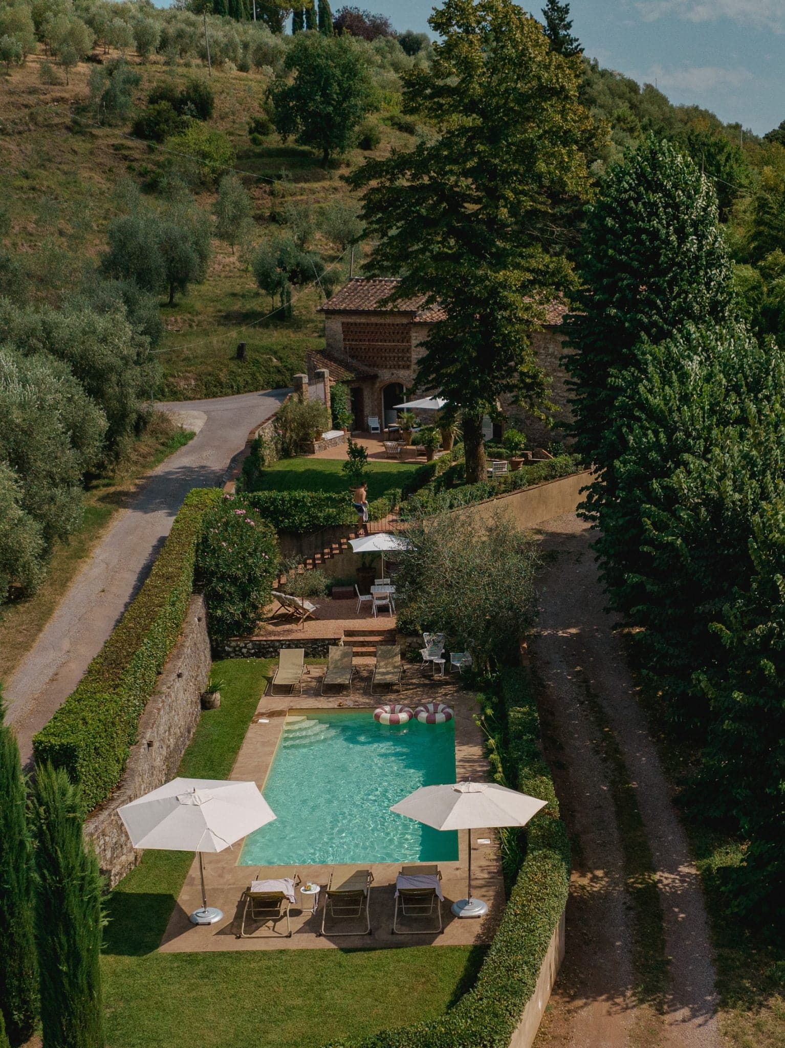 Une scène extérieure sereine avec une piscine rectangulaire entourée d'une végétation luxuriante, avec plusieurs chaises longues et parasols. Un bâtiment rustique est visible en arrière-plan et un chemin serpente à travers l'espace paysager. Des arbres et des collines encadrent le décor.