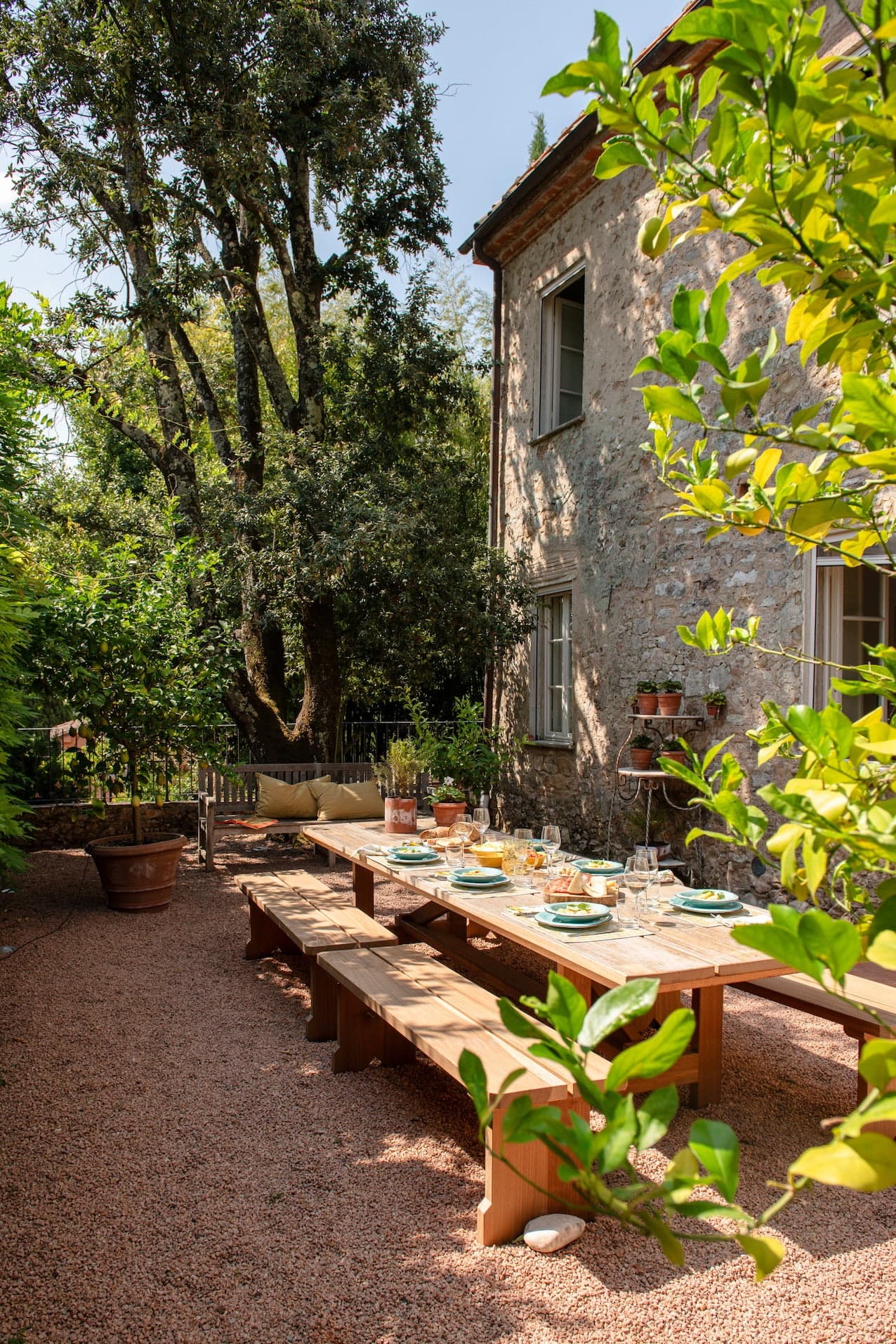 Un espace repas extérieur rustique comprend une longue table en bois dressée pour un repas avec des assiettes, des verres et des couverts. La table, flanquée de bancs en bois, est située sur un patio en gravier à côté d'une vieille maison en pierre. Un feuillage vert luxuriant et un grand arbre entourent le cadre paisible.