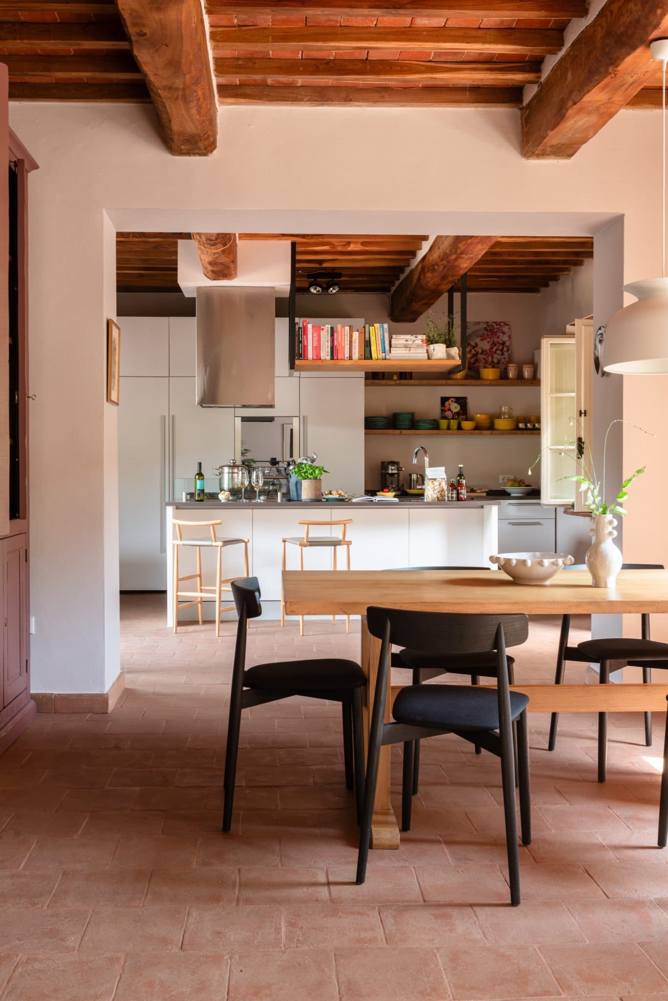 Une cuisine rustique avec des poutres en bois apparentes au plafond, un sol en brique et un mélange de meubles modernes et vintage. Un îlot de cuisine est surmonté de divers ustensiles de cuisine et de deux hauts tabourets en bois. Au premier plan se trouve une table à manger en bois avec des chaises noires.