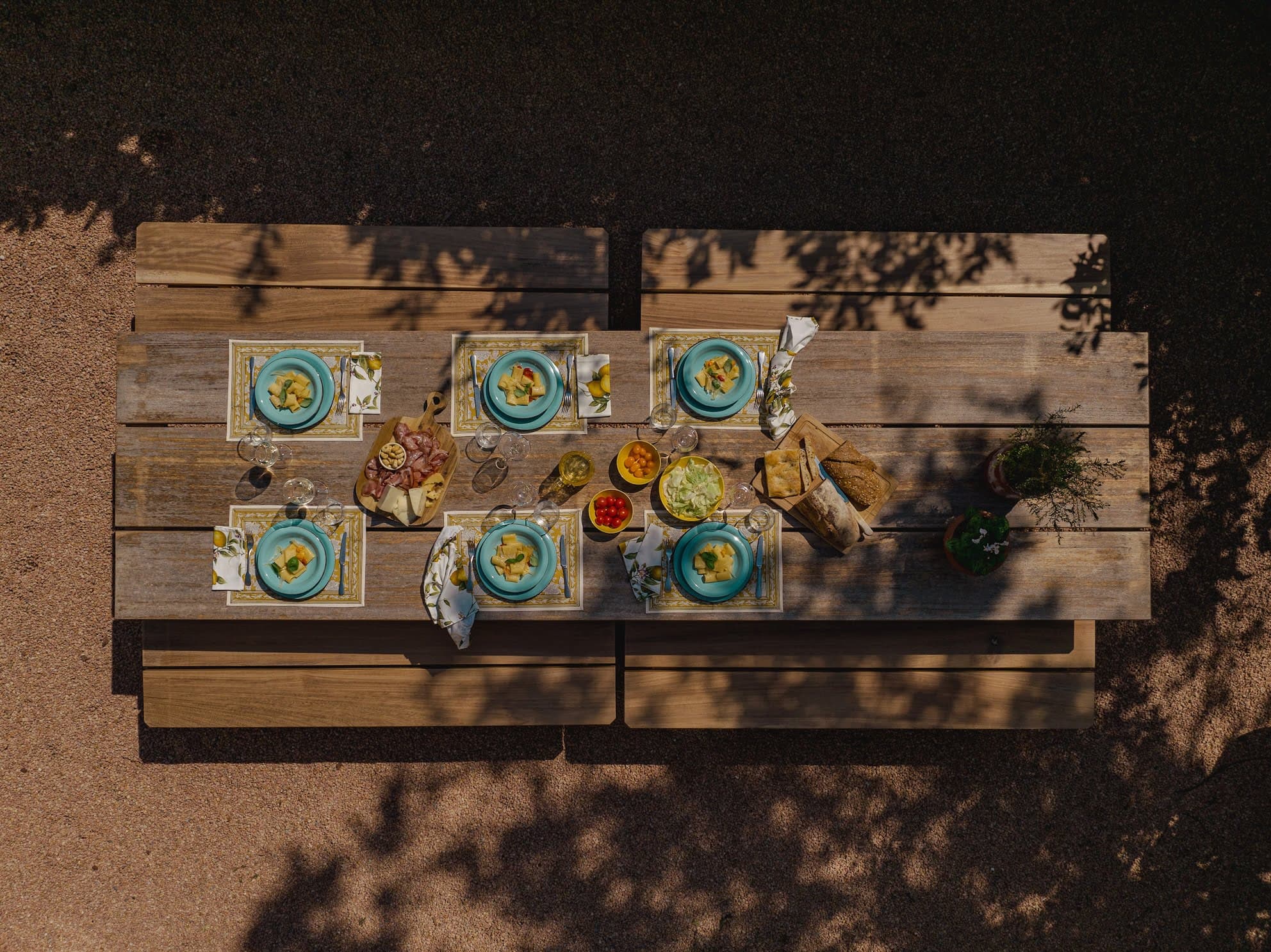 Vue aérienne d'une table de pique-nique en bois dressée pour un repas avec six assiettes bleues remplies de soupe, entourées de divers plats, dont du pain, de la charcuterie, des légumes et des boissons. La table est sous un arbre, projetant des ombres sur elle.