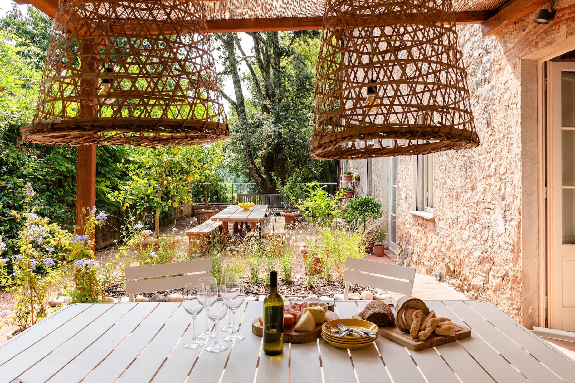 Un patio extérieur confortable avec une table en bois dressée avec du vin, des verres, du fromage et du pain. Des luminaires rustiques en osier sont suspendus au-dessus. L'espace est entouré de verdure luxuriante, de fleurs et d'une longue table à manger de style banc en arrière-plan.