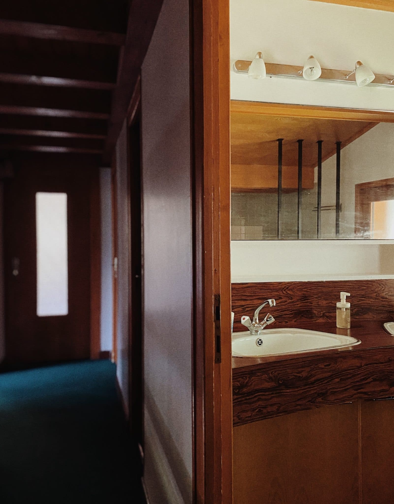 Un couloir faiblement éclairé avec des poutres en bois au plafond mène à une salle de bains. Le comptoir de la salle de bains comprend un lavabo simple avec un robinet moderne, un distributeur de savon et un grand miroir. Au-dessus du miroir, il y a quatre ampoules éteintes.