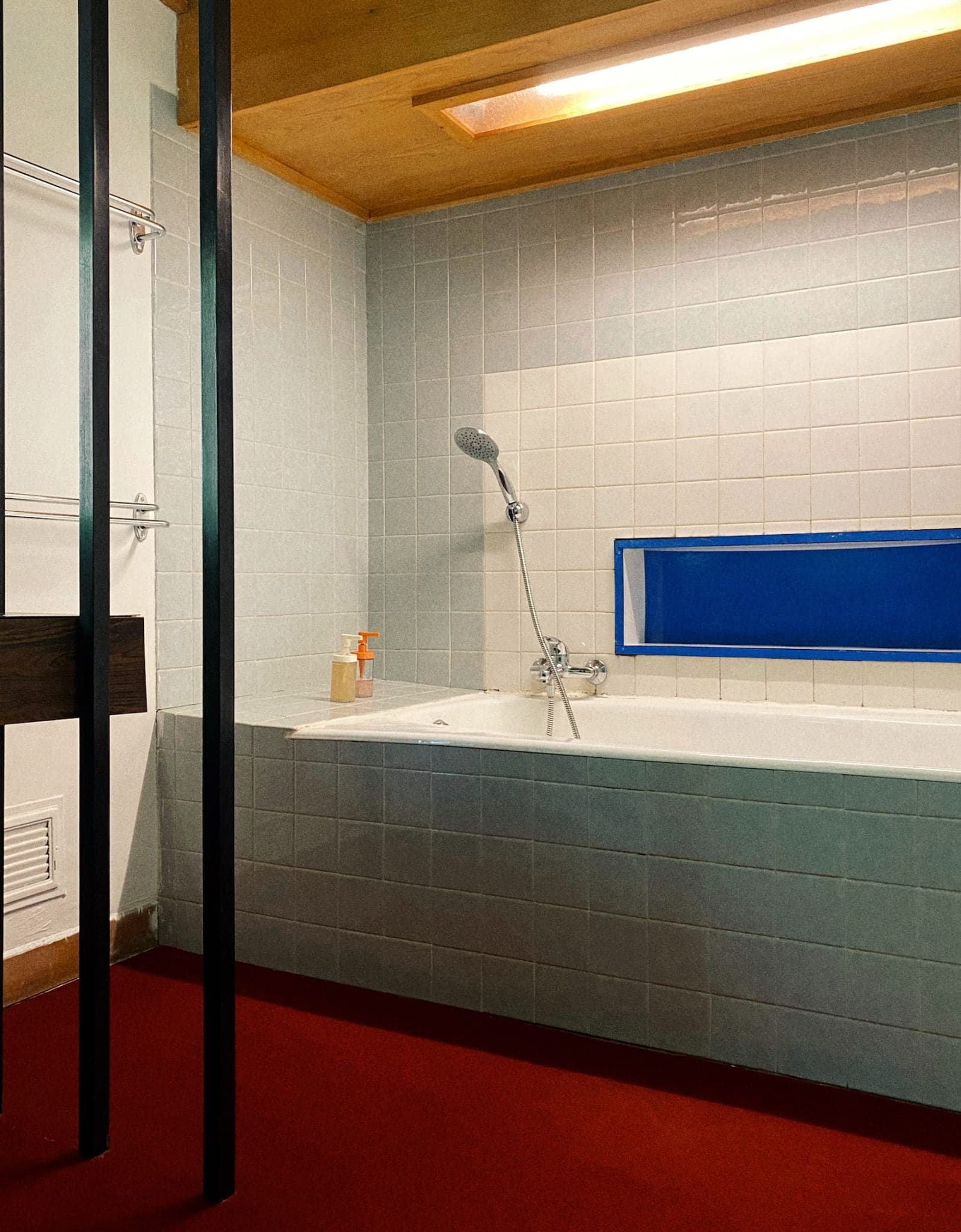 Une salle de bains moderne comprend une grande baignoire blanche aux murs carrelés, une pomme de douche à main et un distributeur de savon. Le sol est recouvert de moquette rouge et des lattes de bois sont utilisées comme luminaires contre le mur. Un éclairage doux et chaleureux illumine l'espace.