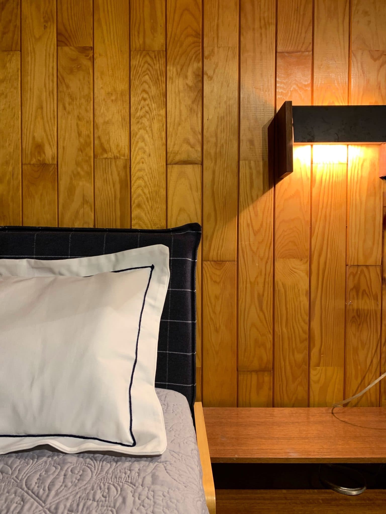 Une chambre confortable avec un mur lambrissé de bois, une tête de lit tapissée de couleur sombre et un lit avec un oreiller blanc avec un passepoil foncé. Une couverture grise matelassée recouvre le lit. Une lampe murale est allumée, émettant une lueur chaleureuse à côté de la tête de lit.