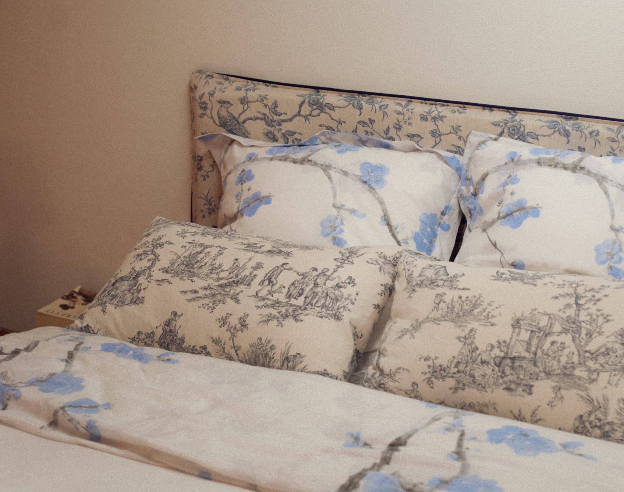 Un lit avec des oreillers et une couverture à motifs floraux. La tête de lit et le couvre-lit présentent des motifs assortis avec des fleurs bleues et grises et des scènes pastorales complexes en noir et blanc. Le mur derrière le lit est de couleur claire.