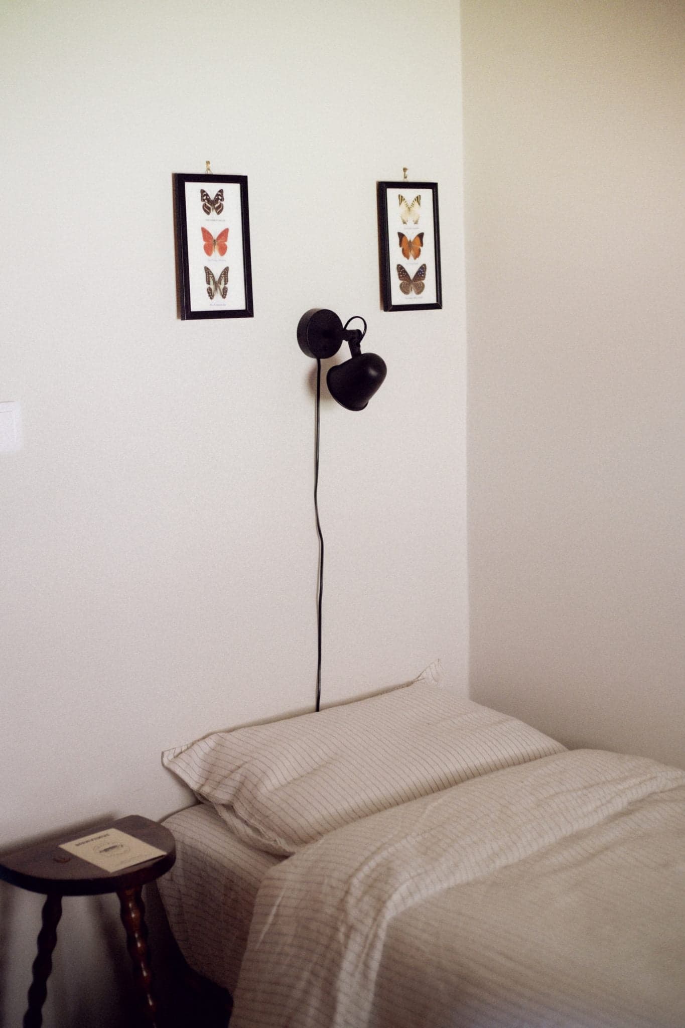 Un lit bien fait avec une literie rayée dans une chambre minimaliste. Au-dessus du lit, deux tableaux encadrés de papillons sont accrochés au mur. Une lampe murale noire avec un cordon est suspendue entre les tableaux. Une petite table d'appoint en bois à côté du lit abrite un livre.