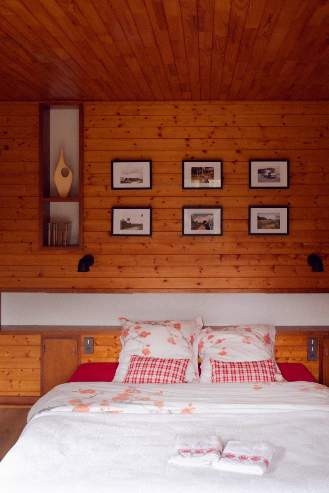 Une chambre confortable avec des murs et un plafond en bois, un lit bien fait avec des oreillers et un couvre-lit blancs et rouges, et cinq tableaux encadrés suspendus au-dessus du lit. Une petite étagère avec un vase et des livres est encastrée dans le mur à gauche. Des serviettes pliées reposent sur le lit.