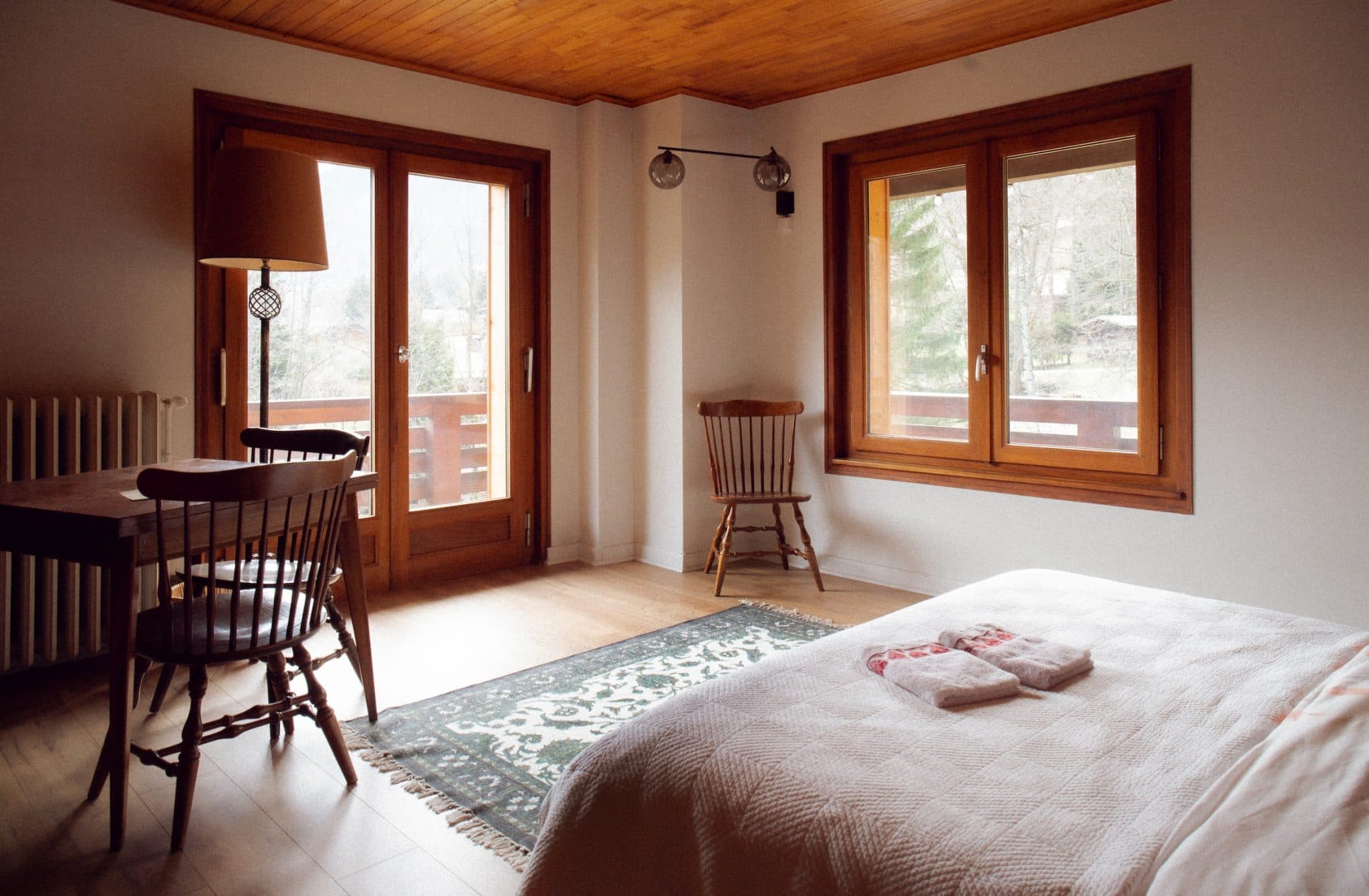 Une chambre confortable avec des meubles en bois comprend un lit avec des serviettes pliées dessus, un tapis sur un plancher en bois, une table et des chaises près d'un radiateur et de grandes fenêtres qui laissent entrer la lumière naturelle, offrant une vue sur les arbres à l'extérieur. Un plafonnier est suspendu au-dessus.