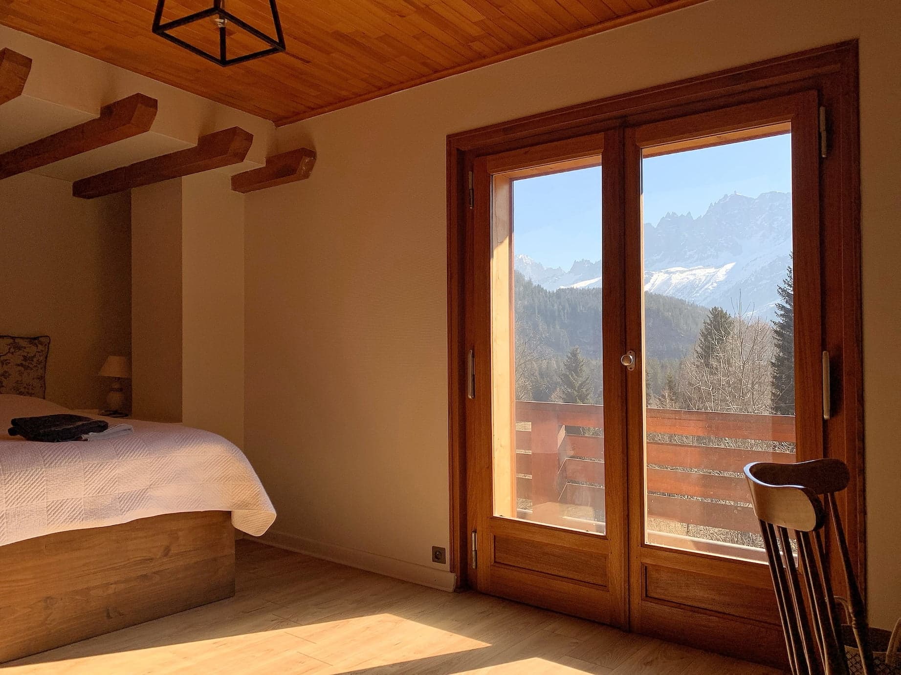 Une chambre sereine avec plafond en bois et poutres apparentes, et un grand lit recouvert d'une couverture blanche. La lumière du soleil pénètre à travers une porte vitrée qui s'ouvre sur un balcon avec vue sur la montagne et la forêt. Une chaise en bois est installée dans un coin et des vêtements pliés reposent sur le lit.