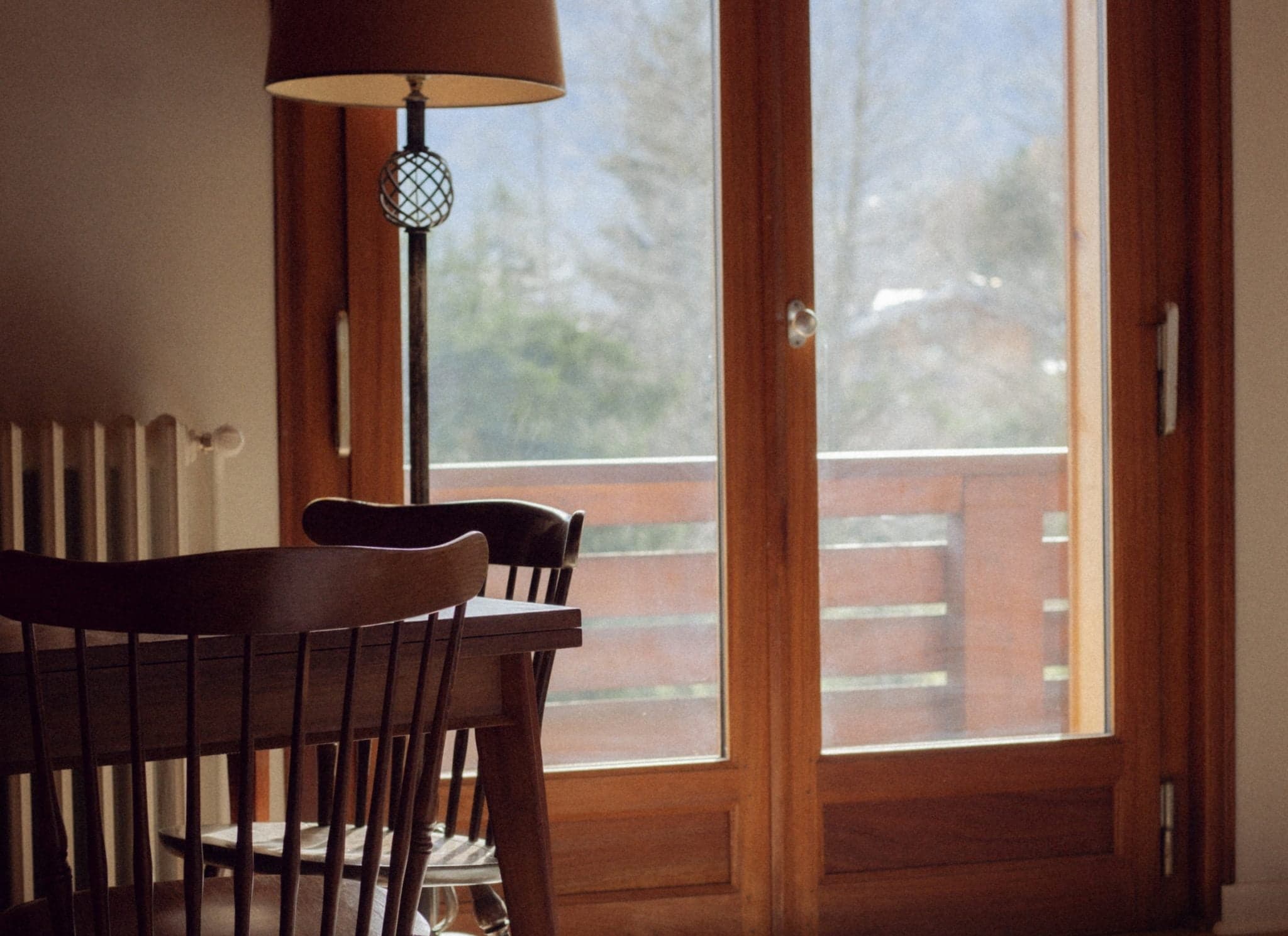 Une table à manger en bois confortable et des chaises sont placées près d'une grande fenêtre avec vue sur un balcon et des arbres au loin. Un lampadaire se trouve à côté de la table, projetant une lumière chaleureuse, renforçant l'atmosphère sereine et accueillante de la pièce.