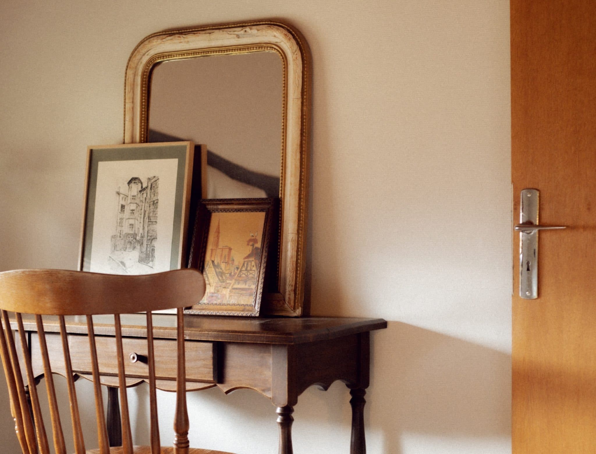 Une chaise en bois est placée devant un bureau en bois. Sur le bureau, un grand miroir décoré repose contre le mur, avec deux tableaux encadrés posés devant lui. Une porte en bois avec une poignée en métal est partiellement visible sur le côté droit de l'image.