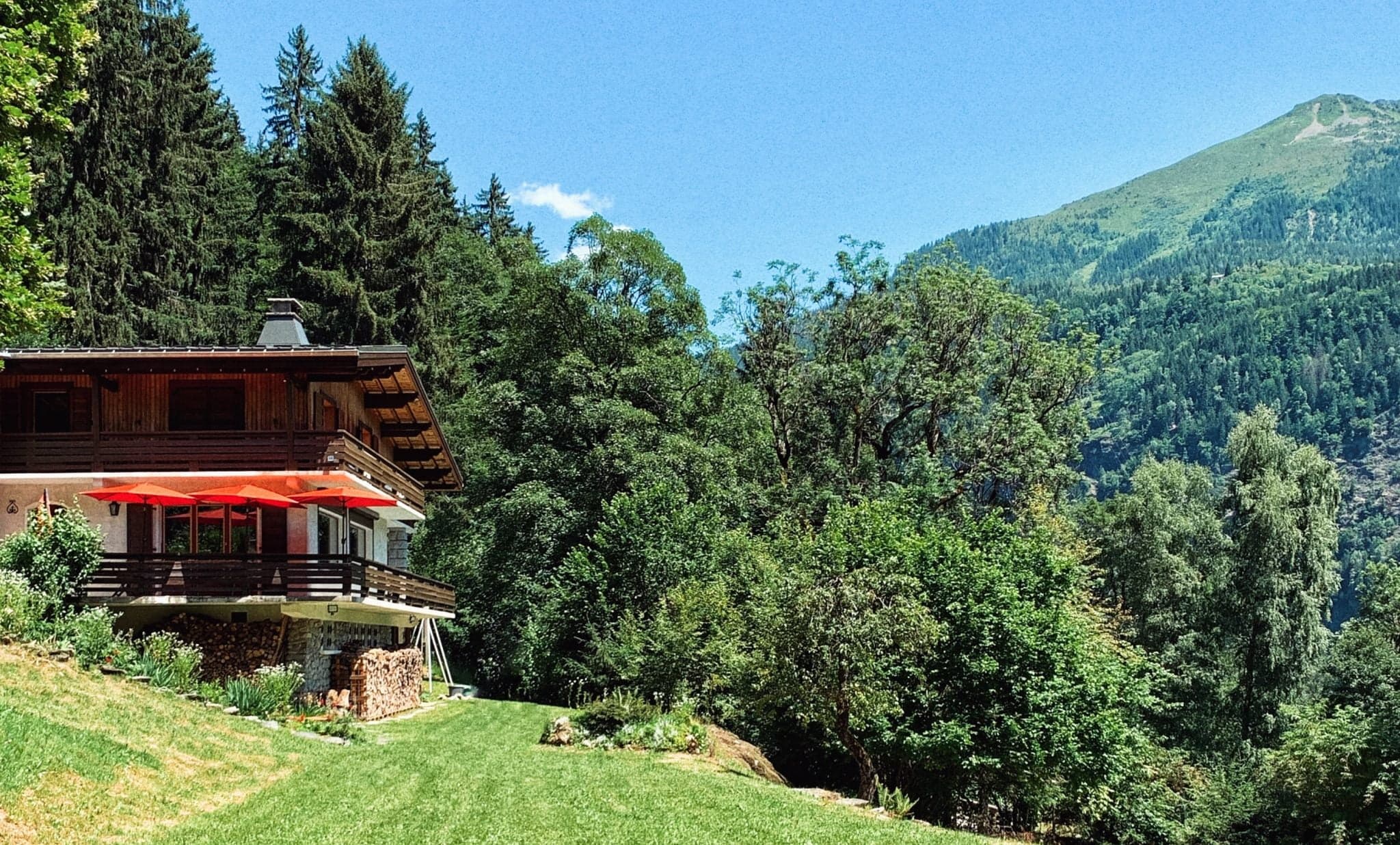 A charming wooden cottage with red umbrellas on the deck sits on a vibrant green lawn, surrounded by dense trees. The backdrop features a lush mountain range under a clear blue sky.