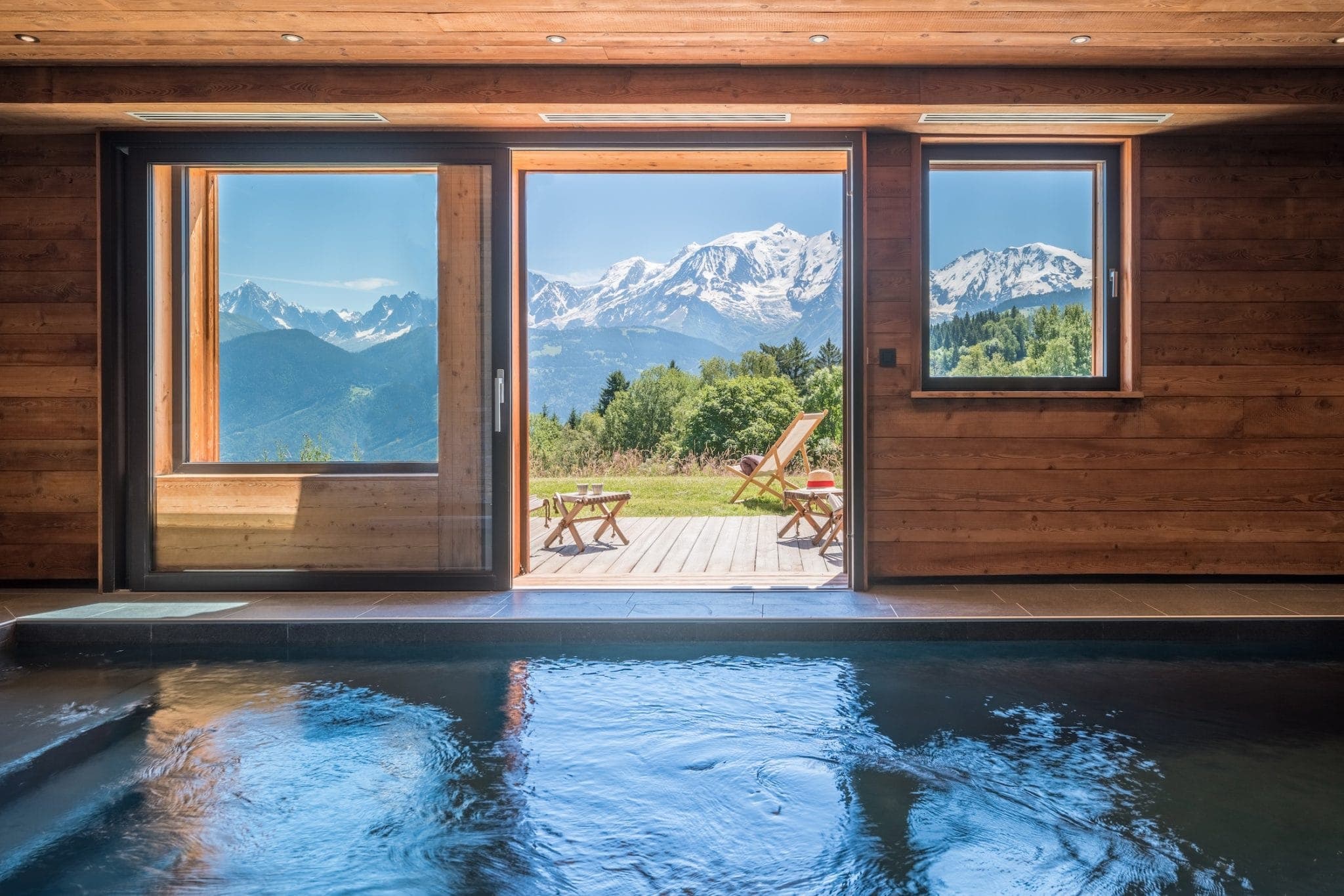 An indoor swimming pool with a view of snowy mountains through large glass doors and windows. Outside, there are wooden chairs on a deck. The interior features wooden walls and a serene atmosphere, blending indoor comfort with breathtaking natural scenery.