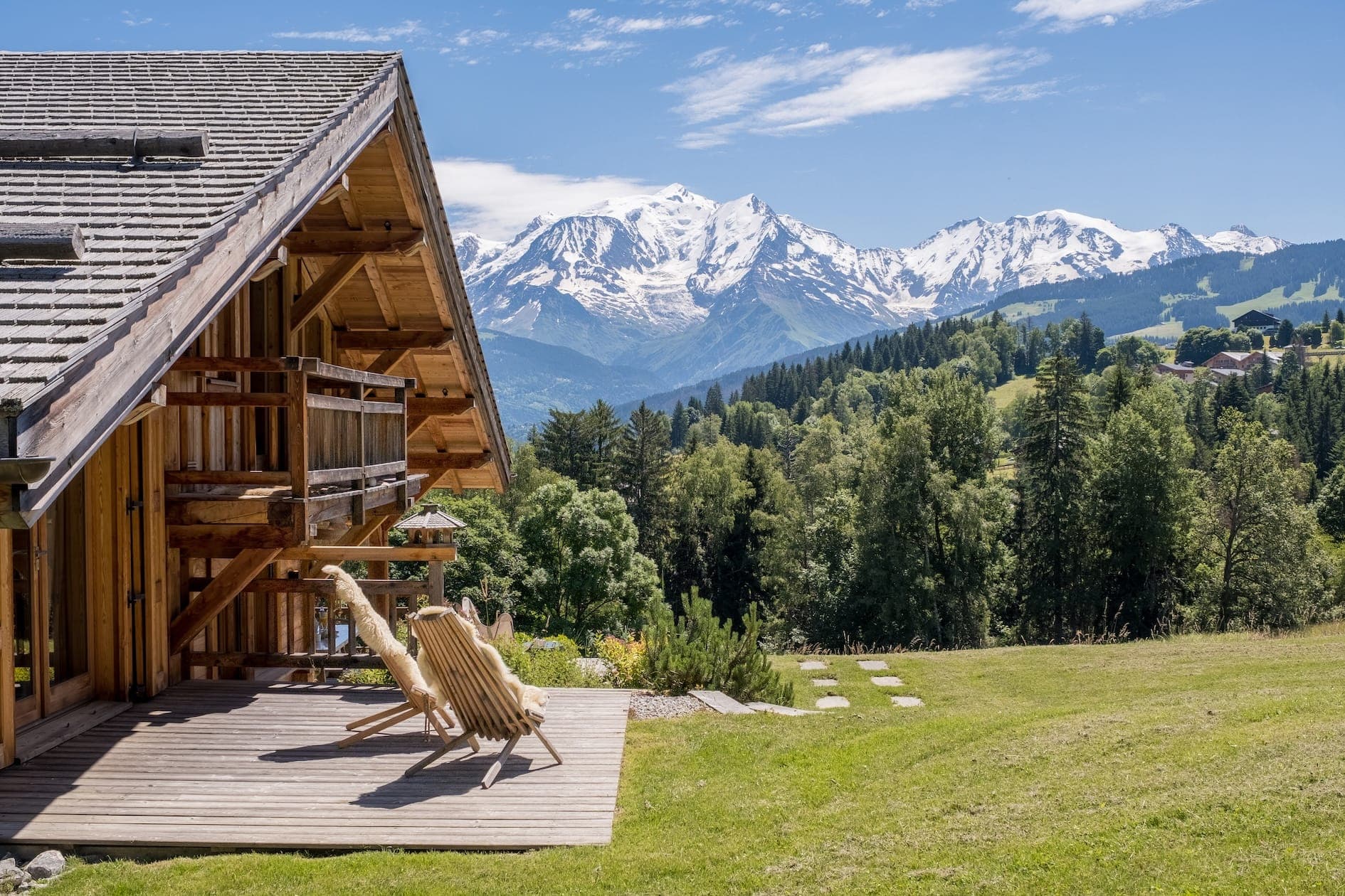 Un chalet de montagne pittoresque avec une terrasse en bois et deux chaises longues. Le chalet est entouré d'arbres verts luxuriants et d'herbe, avec en toile de fond un magnifique paysage de sommets enneigés sous un ciel bleu.