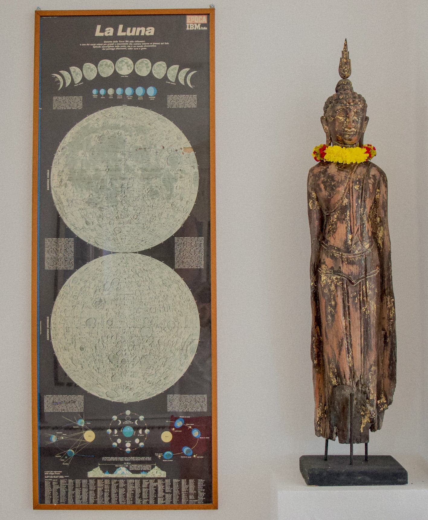 Une affiche encadrée représentant différentes phases lunaires et des cartes de la lune est accrochée sur un mur blanc à côté d'une grande statue patinée par les intempéries d'un Bouddha debout orné d'une guirlande de fleurs jaunes et rouges autour du cou. La statue est sur un piédestal blanc.