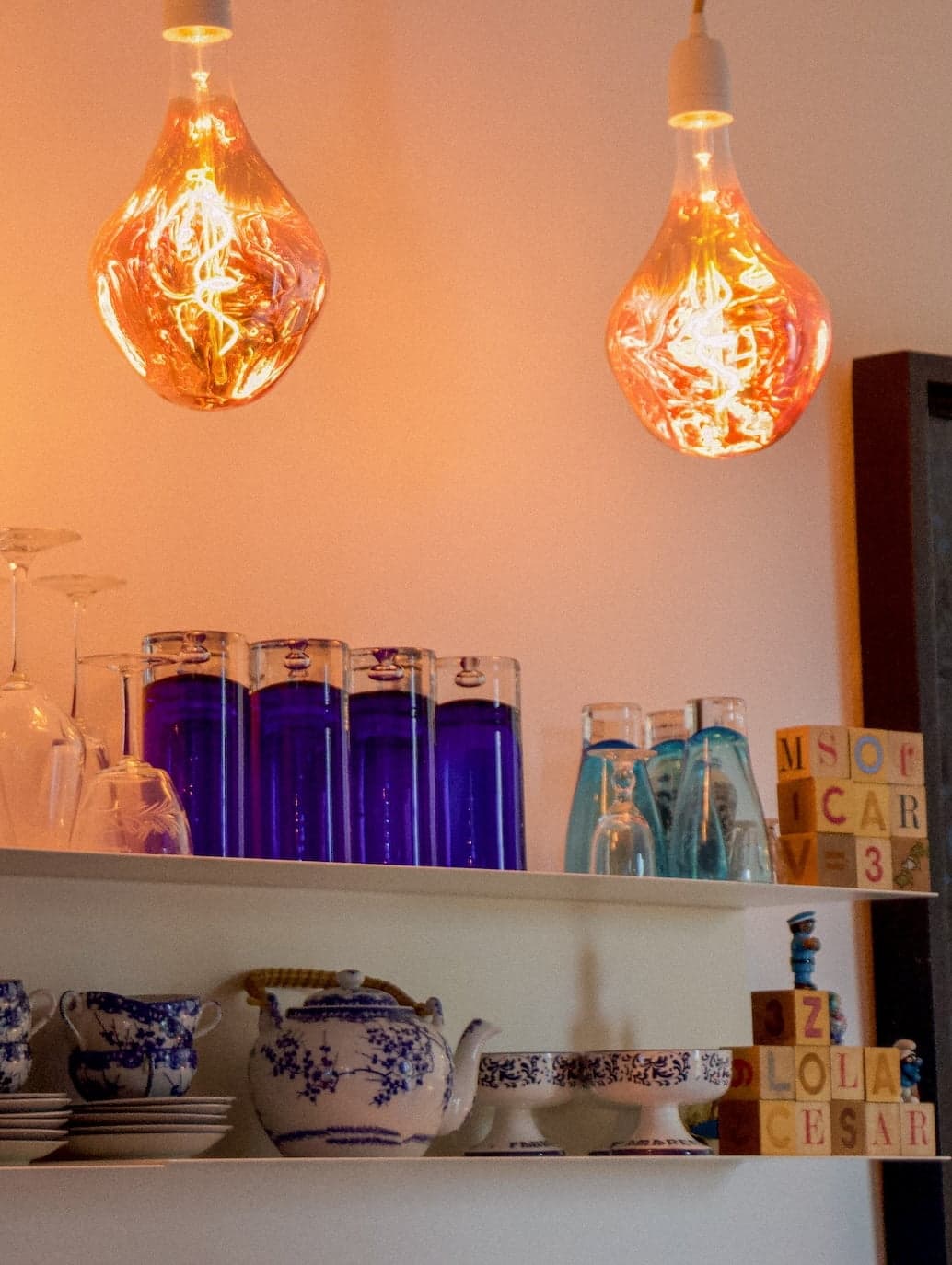 Une étagère sur laquelle sont exposés divers objets : des verres à boire teintés de bleu, des plats en céramique bleu et blanc aux motifs complexes et des piles de blocs de lettres en bois. Au-dessus de l'étagère, deux lampes suspendues avec des ampoules à filament décoratives émettent une lueur chaleureuse.