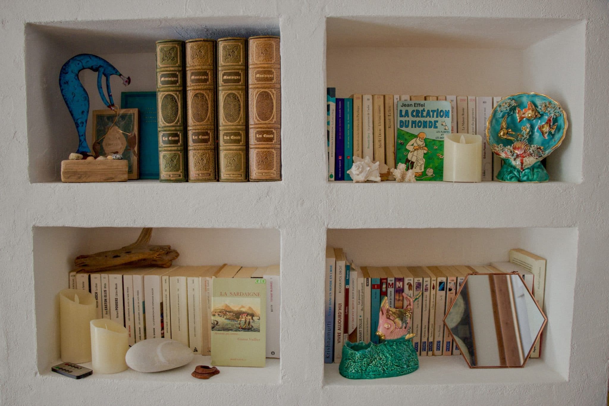 Deux étagères murales accueillent des livres et des objets de décoration. L'étagère supérieure accueille un mélange de livres, deux coquillages, une bougie blanche et une décoration sur le thème des papillons. L'étagère inférieure abrite des livres, une sculpture de visage rose en céramique verte et un miroir hexagonal.