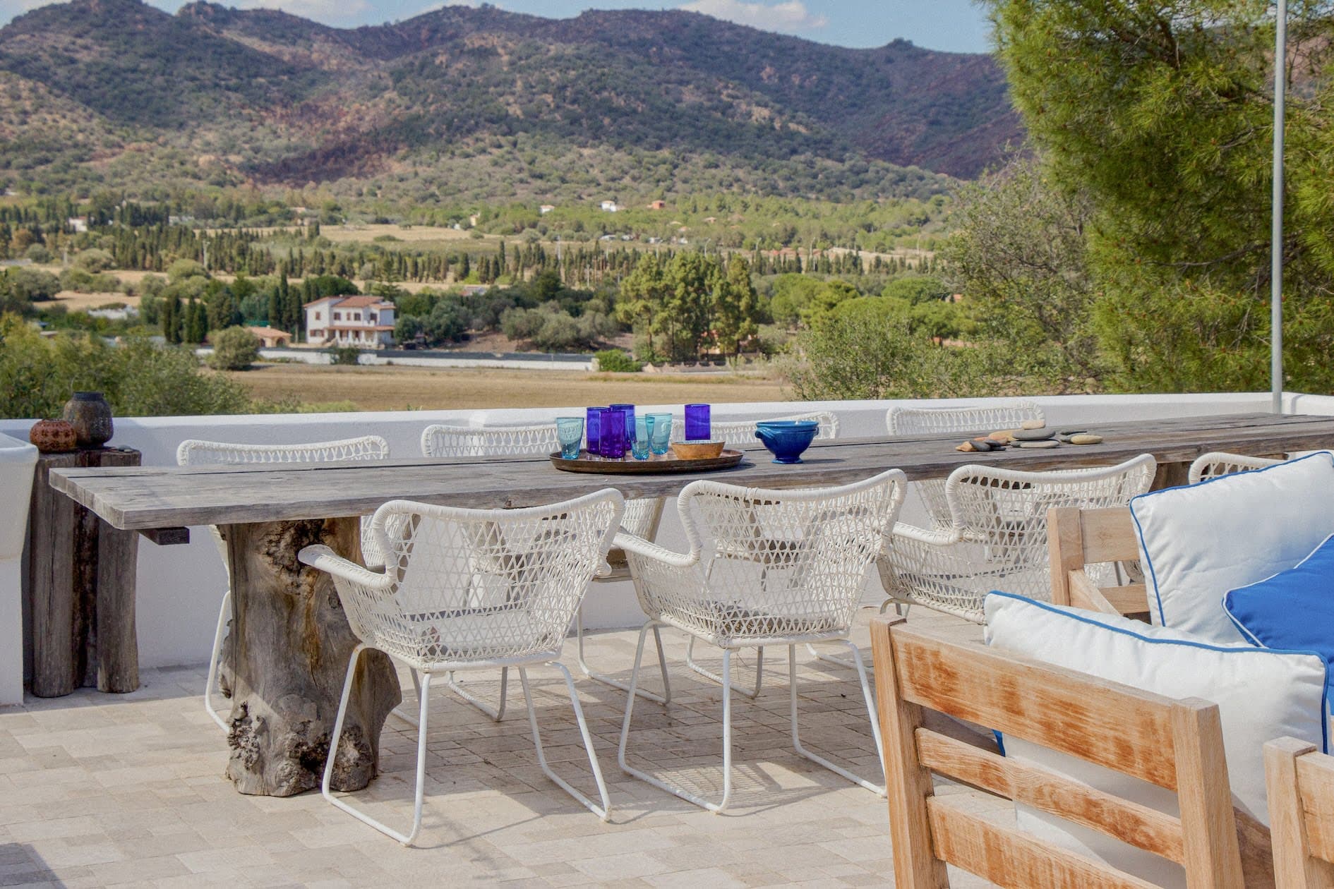 Un espace repas extérieur avec une table en bois rustique entourée de chaises en osier blanc. La table est dressée avec des verres colorés. L'espace donne sur une vue panoramique sur les montagnes et la verdure sous un ciel partiellement nuageux. À proximité, un banc en bois rembourré est visible.