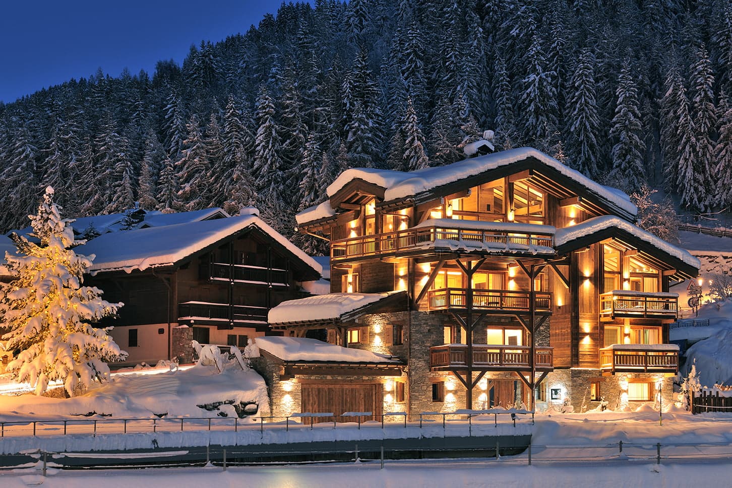 Un chalet en bois à plusieurs étages, chaleureusement éclairé et doté de larges baies vitrées, se niche au milieu des conifères enneigés au crépuscule. Les balcons et le toit du chalet sont chargés de neige, ce qui donne une atmosphère chaleureuse et accueillante dans un ciel crépusculaire. Un autre bâtiment en bois plus petit se trouve à proximité.