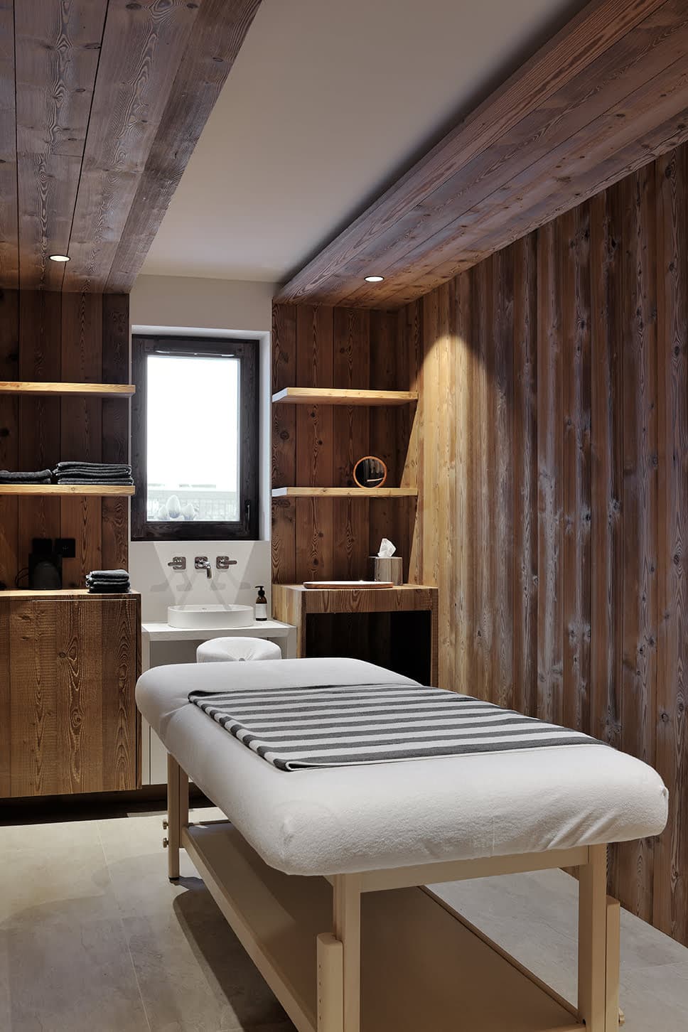 A modern and serene spa treatment room featuring wooden walls and ceiling. A neatly made massage table with a striped towel is centered in the room. Shelves with decor items, including towels and a small potted plant, are on the walls. Natural light filters in through a window.