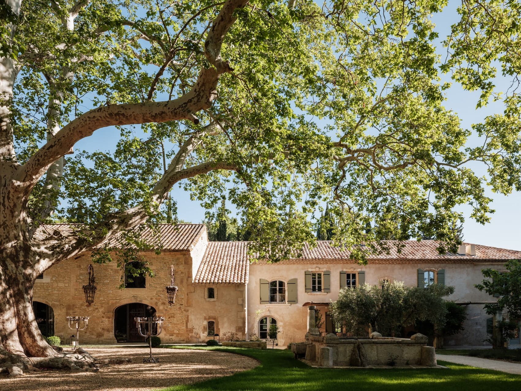 A serene scene of a rustic estate with a tan stone façade, red-tiled roof, and green shutters. Tall, leafy trees with sprawling branches shade the grassy yard, where a decorative fountain and wrought-iron candelabra add charm to the tranquil setting.