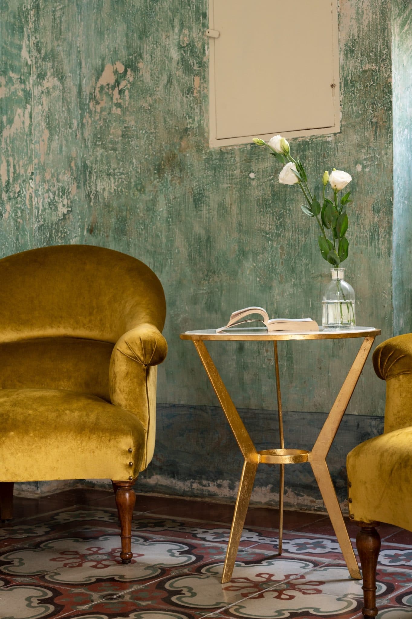 A serene corner features two mustard velvet armchairs, a small table with triangular legs topped with a glass vase containing white flowers and an open book. The background includes a textured green wall and patterned tile flooring. Natural light filters through a window.