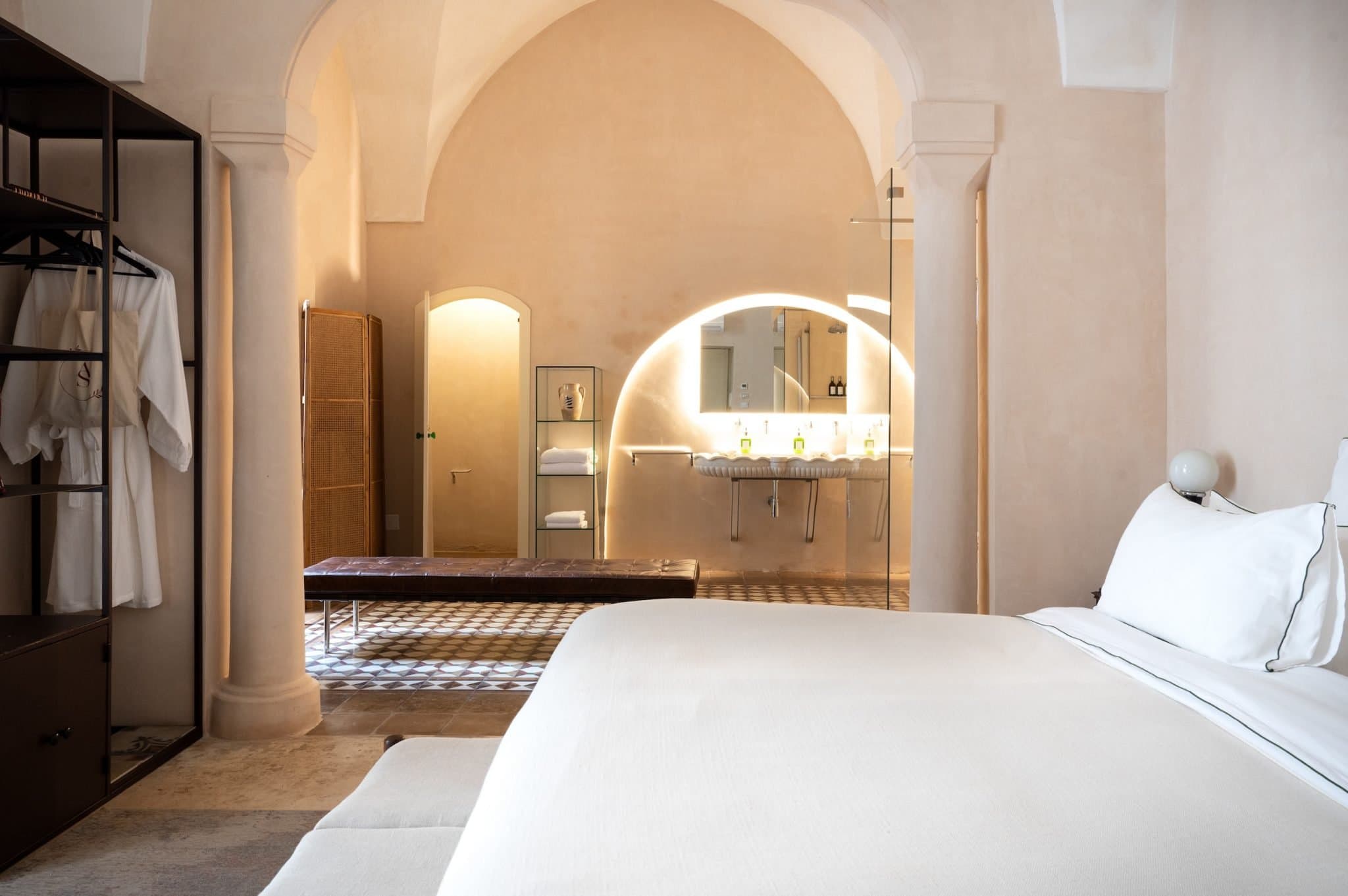A luxurious and minimalist hotel room features a neatly made bed with white linens, an open shelf with bathrobes, and a view of a modern, well-lit bathroom with an arched mirror and double sink. The room has high ceilings and soft, neutral tones.