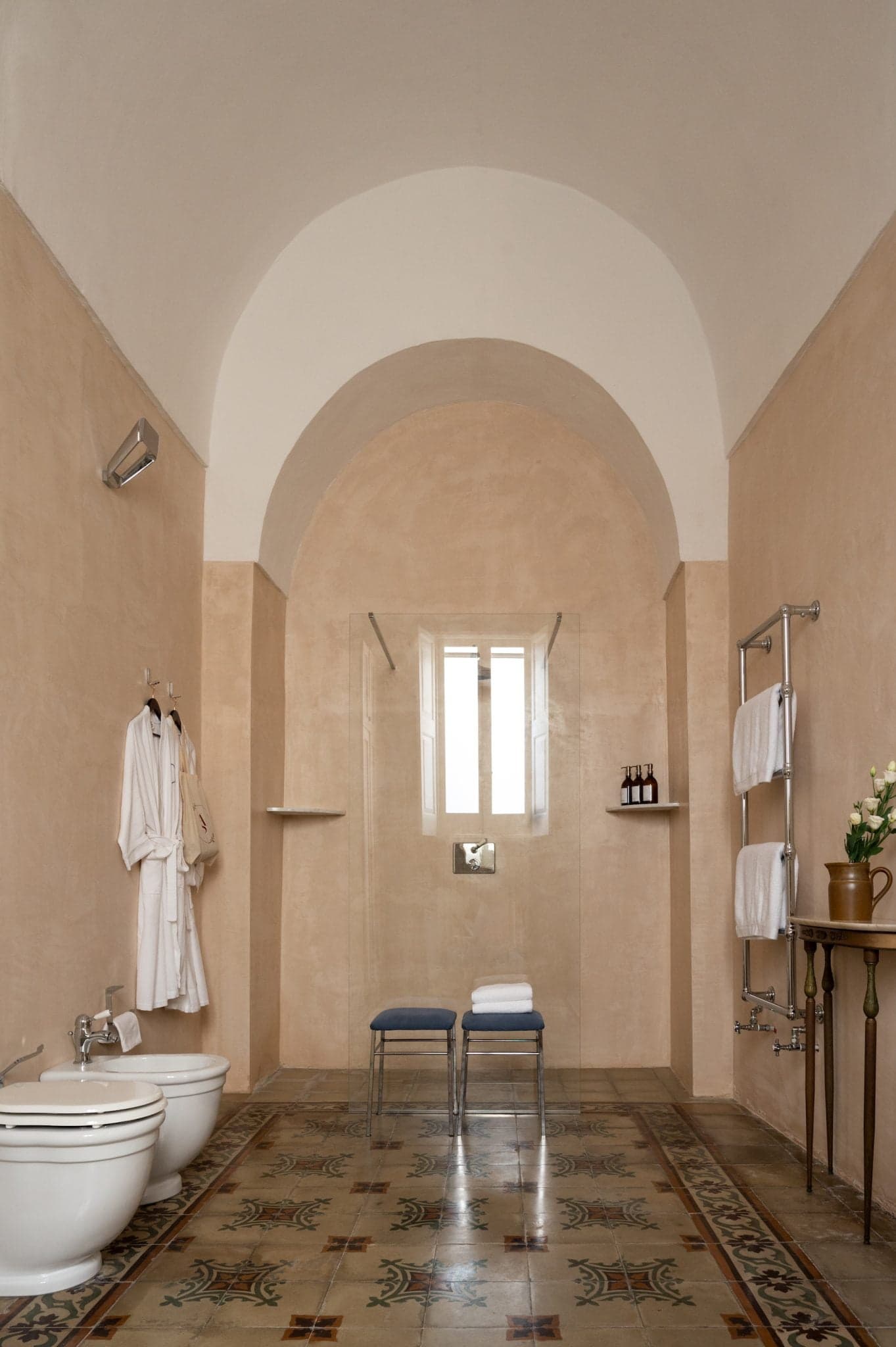 The spacious bathroom includes a shower area with a curved ceiling, flanked by two bidets on the left and a freestanding towel rack with shelves and towels on the right. A small table with flowers and toiletries adds a decorative touch, while bathrobes are hung near the door.