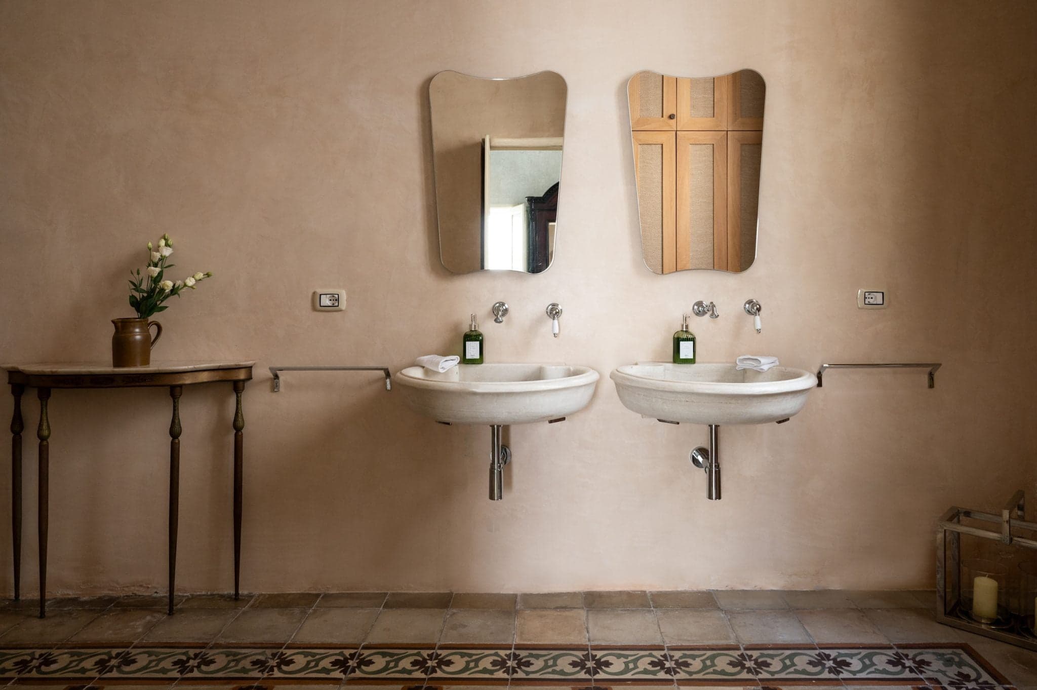 A bathroom with two white sinks beneath irregularly shaped mirrors, each with a green soap dispenser. To the left, there is a small table with a vase of white flowers, and the floor is decorated with patterned tiles. On the right, a wooden panel and a basket with a candle are placed.