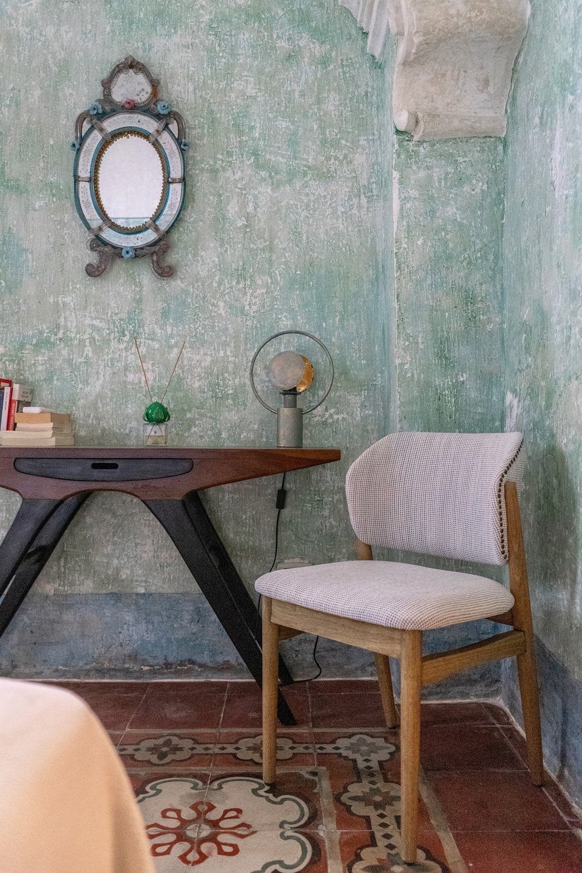 A small room with textured green walls features a wooden table holding books, a diffuser, and a unique lamp. An oval framed mirror hangs above the table. A cushioned chair with light upholstery and wooden legs is placed near the table, which rests on a patterned floor.