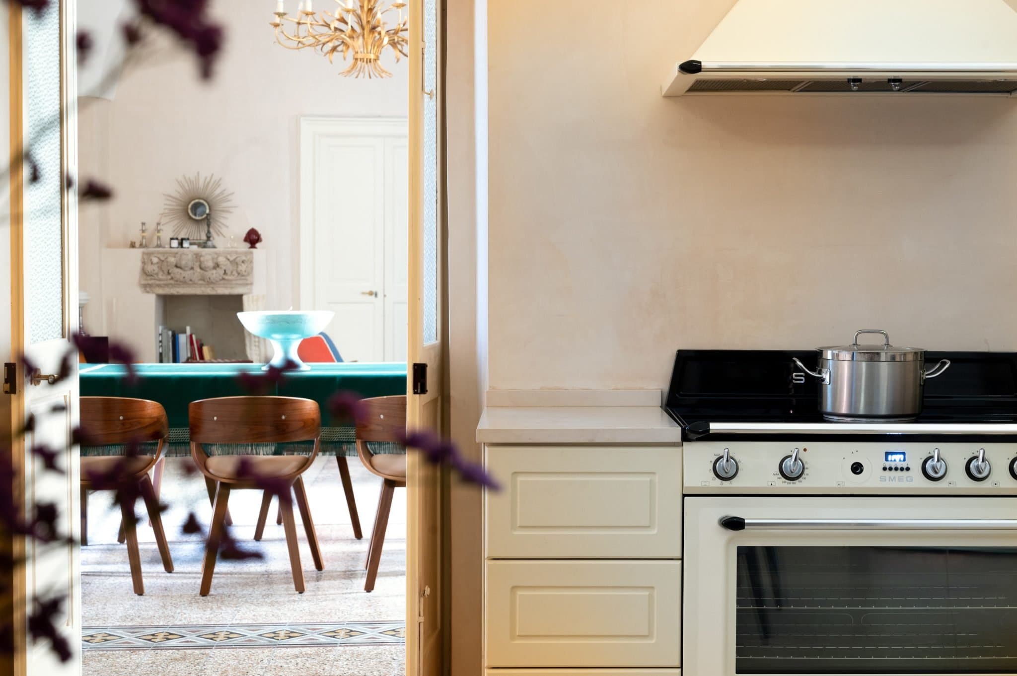 View from a kitchen into a dining room. The kitchen features light-colored cabinets, a black stove with a pot, and a white range hood. The dining room has wooden chairs around a table, a decorative fireplace, a chandelier, and wilted purple flowers in the foreground.