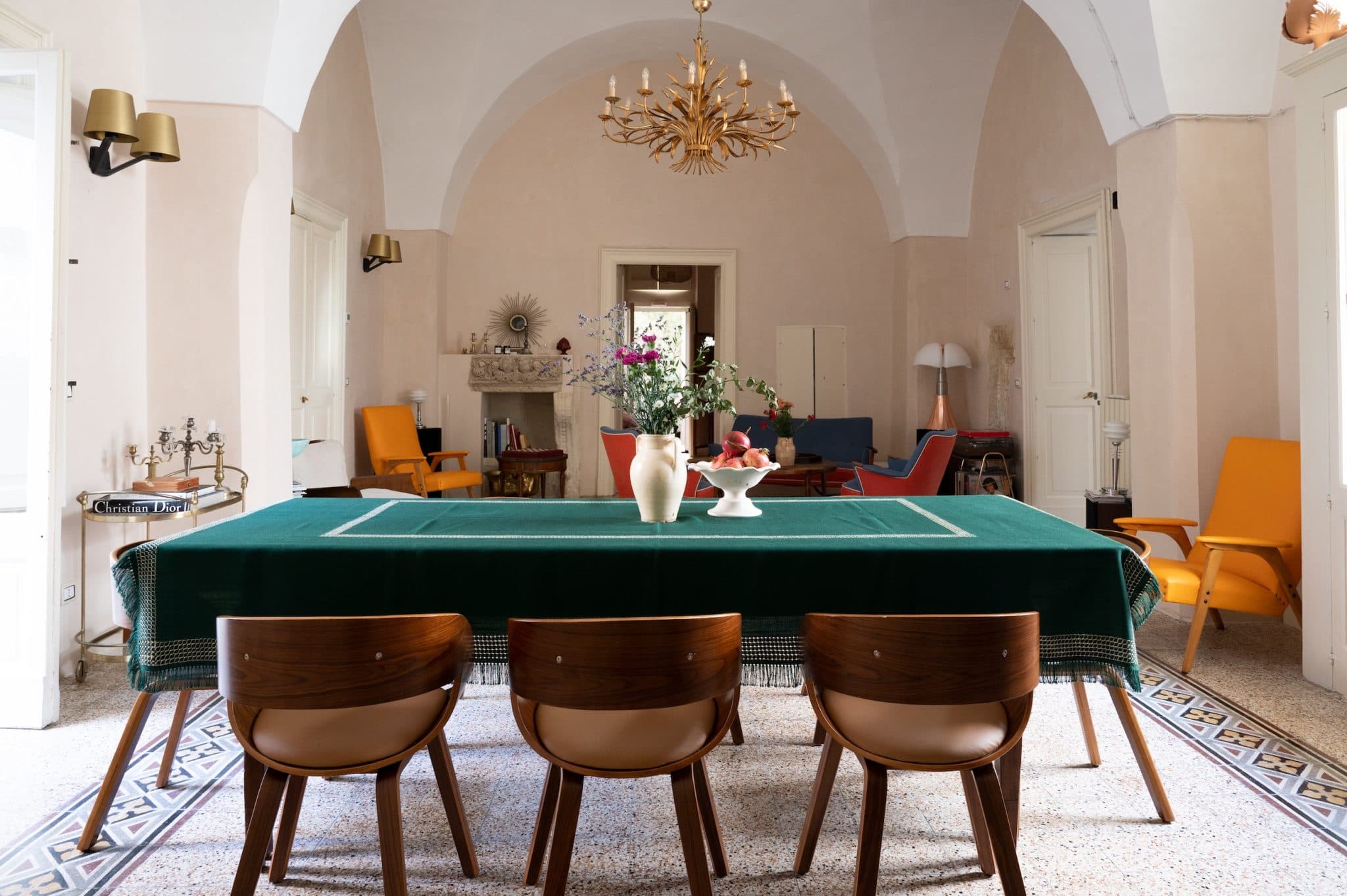 A spacious and elegant dining room features a table covered with a green tablecloth adorned with floral arrangements and white ceramics. Wooden chairs are arranged around the table. The background includes vaulted ceilings, modern orange armchairs, and a chandelier.