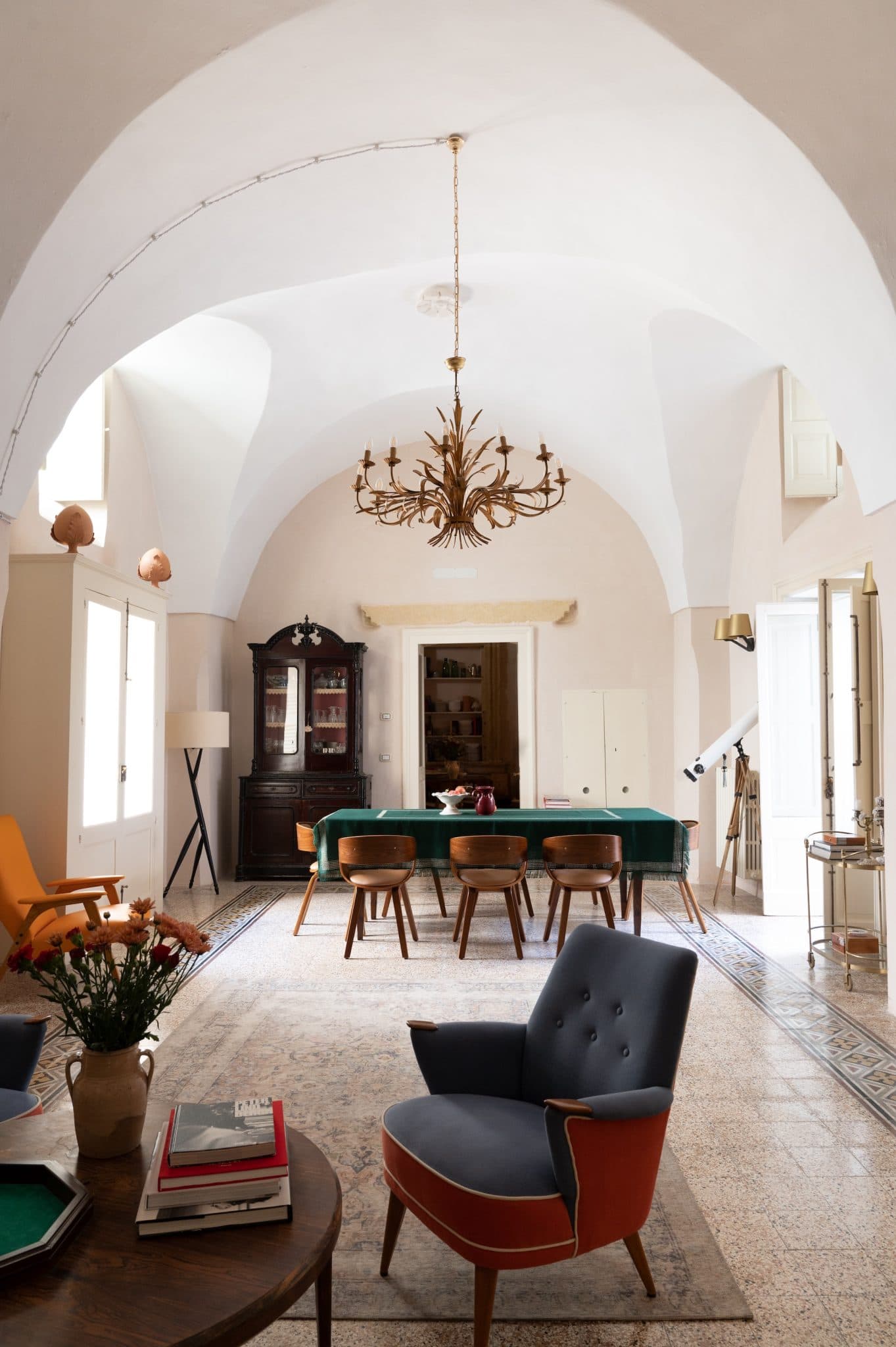 A spacious and elegant room features a large chandelier, a long table with six chairs, a blue retro armchair, and a polished wooden cabinet. The room boasts high vaulted ceilings, light-colored walls, and abundant natural light from the windows. A telescope is situated near the corner.