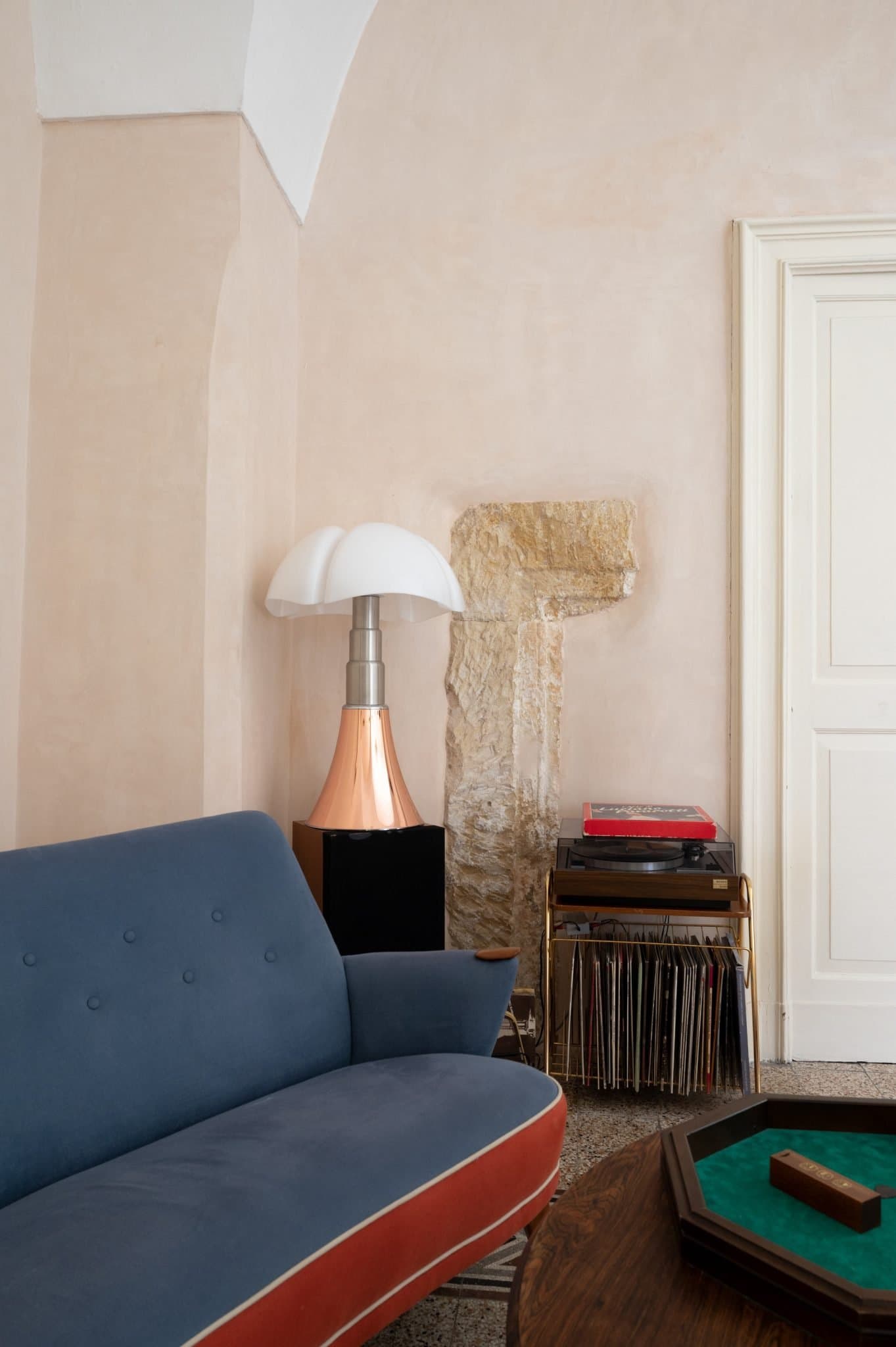 A cozy seating area with a blue and red sofa, a retro lamp with a mushroom-shaped lampshade on a side table, and a collection of vinyl records on a nearby shelf. A white door and a textured beige wall complete the scene, creating a relaxed atmosphere.