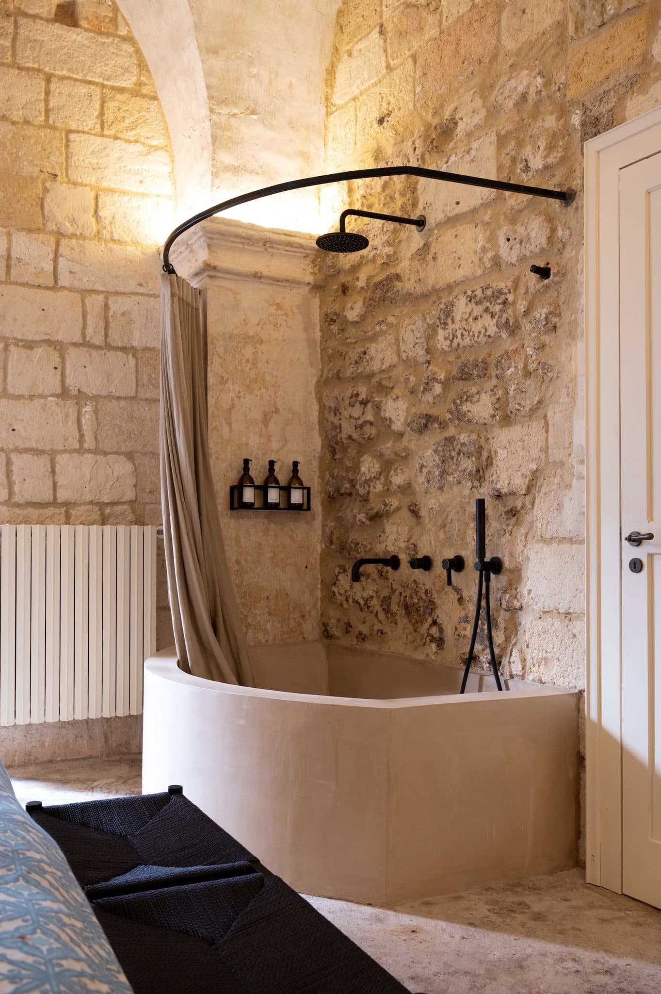 A bathroom with a rustic stone wall in the background features a built-in bathtub with a curved shower rod and curtain. The dark fixtures include a rain showerhead, a faucet, and a mounted shelf that can hold three bottles. The floor is made of stone, and a white door is partially visible on the right.