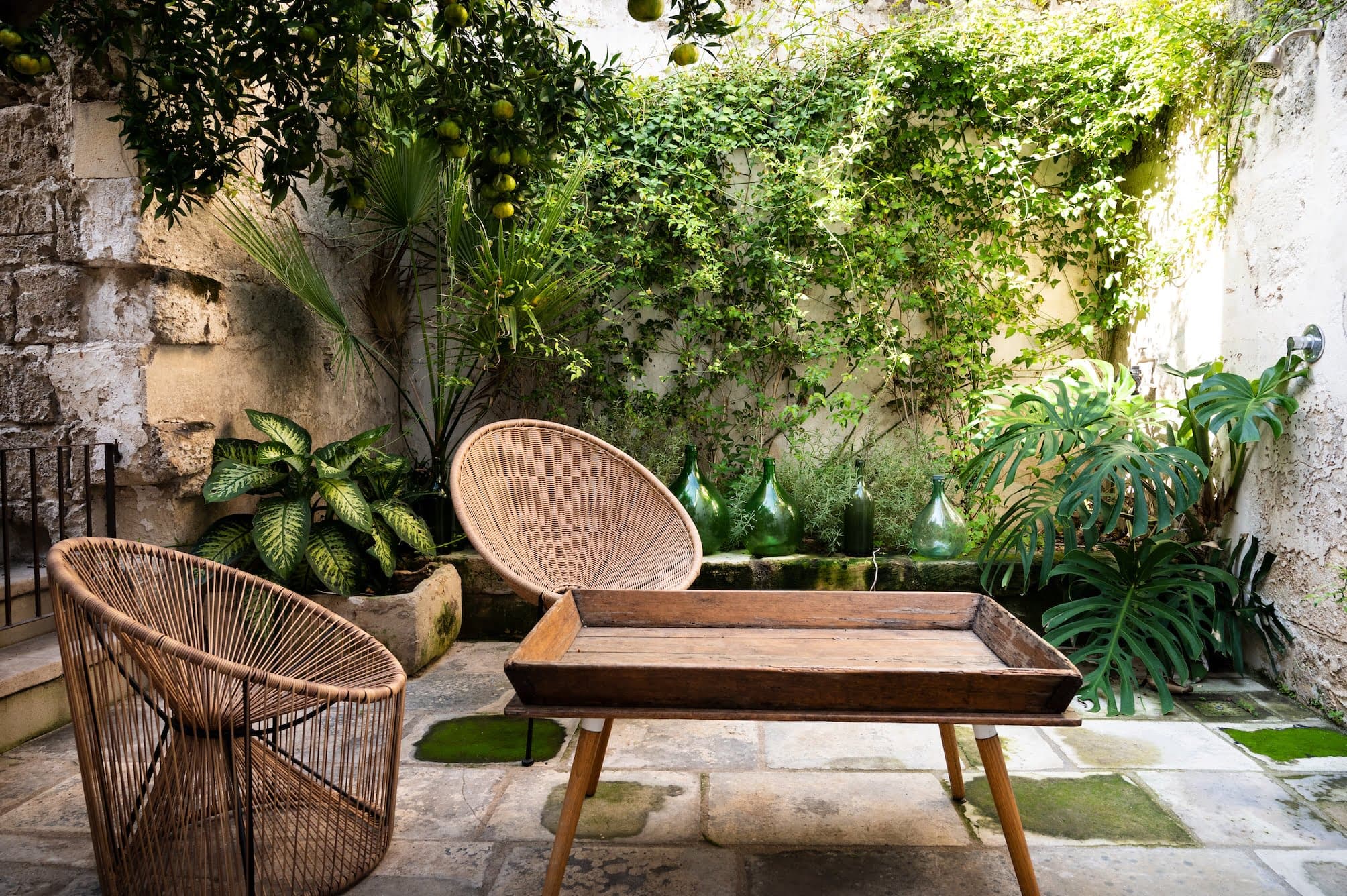 A peaceful outdoor patio with green vines covering a stone wall. Two wicker chairs are placed around a rectangular wooden table. The patio floor is paved with stone tiles, and an assortment of potted plants adds lush greenery to the serene setting.