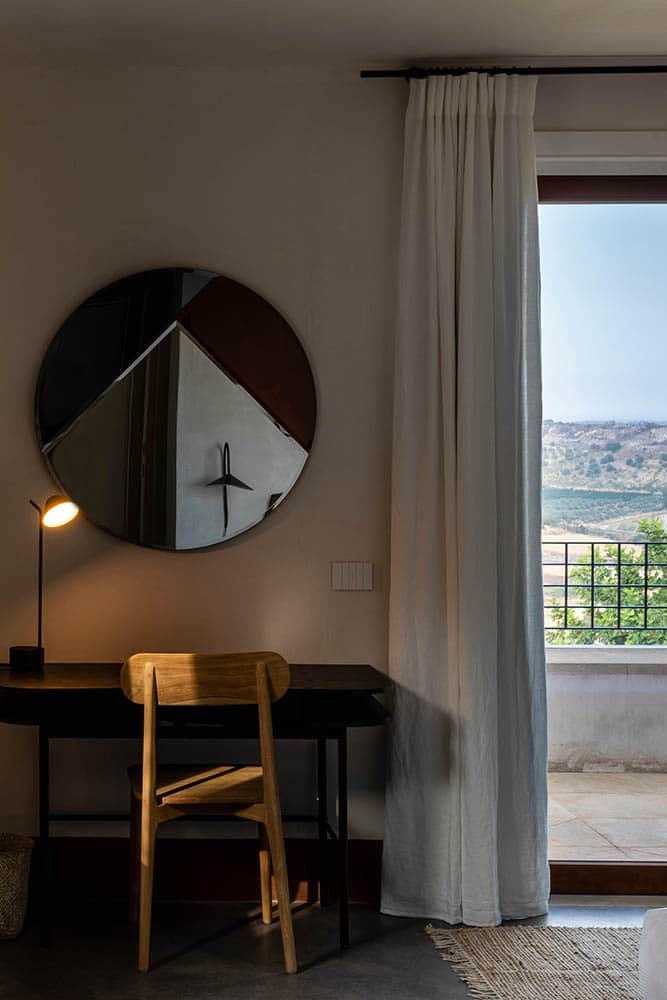 A modern bedroom with a wooden desk and chair, a round mirror hanging above the desk, and a small lamp on it. A large window with partially drawn white curtains reveals a view of an outdoor balcony and distant hills.