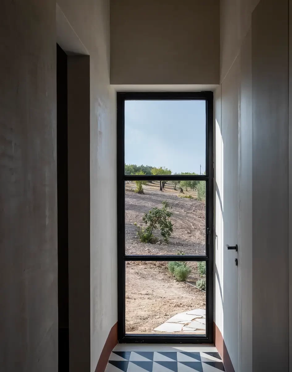 View through a narrow hallway with a glass door at the end. The door opens onto an outdoor scene with sparse, dry vegetation and trees. The hallway features minimalist interiors with a geometric-patterned floor. Natural light streams in abundantly.