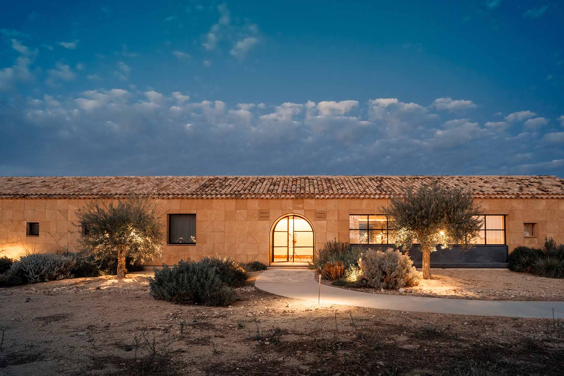 Une maison rustique en pierre de plain-pied avec un toit en tuiles est illuminée au crépuscule. La porte d'entrée brille chaleureusement, entourée d'un paysage serein de plantes du désert et de petits arbres. Le ciel au-dessus est parsemé de nuages doux, créant une atmosphère tranquille.