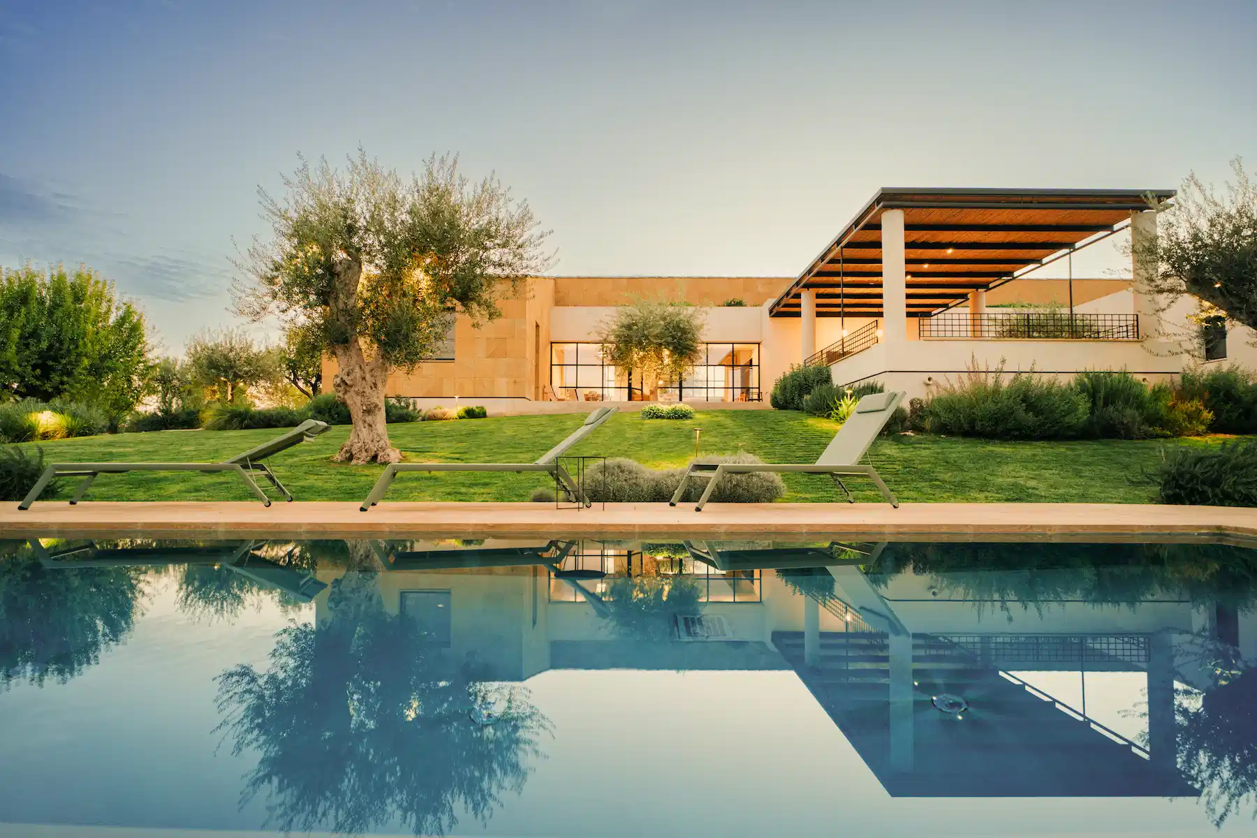 A modern villa set against a clear sky features a spacious courtyard with neatly trimmed grass, several trees, and a pool. Lounge chairs line the edge of the pool, and the villa has large windows and a shaded patio. The calm water of the pool reflects the serene scene.