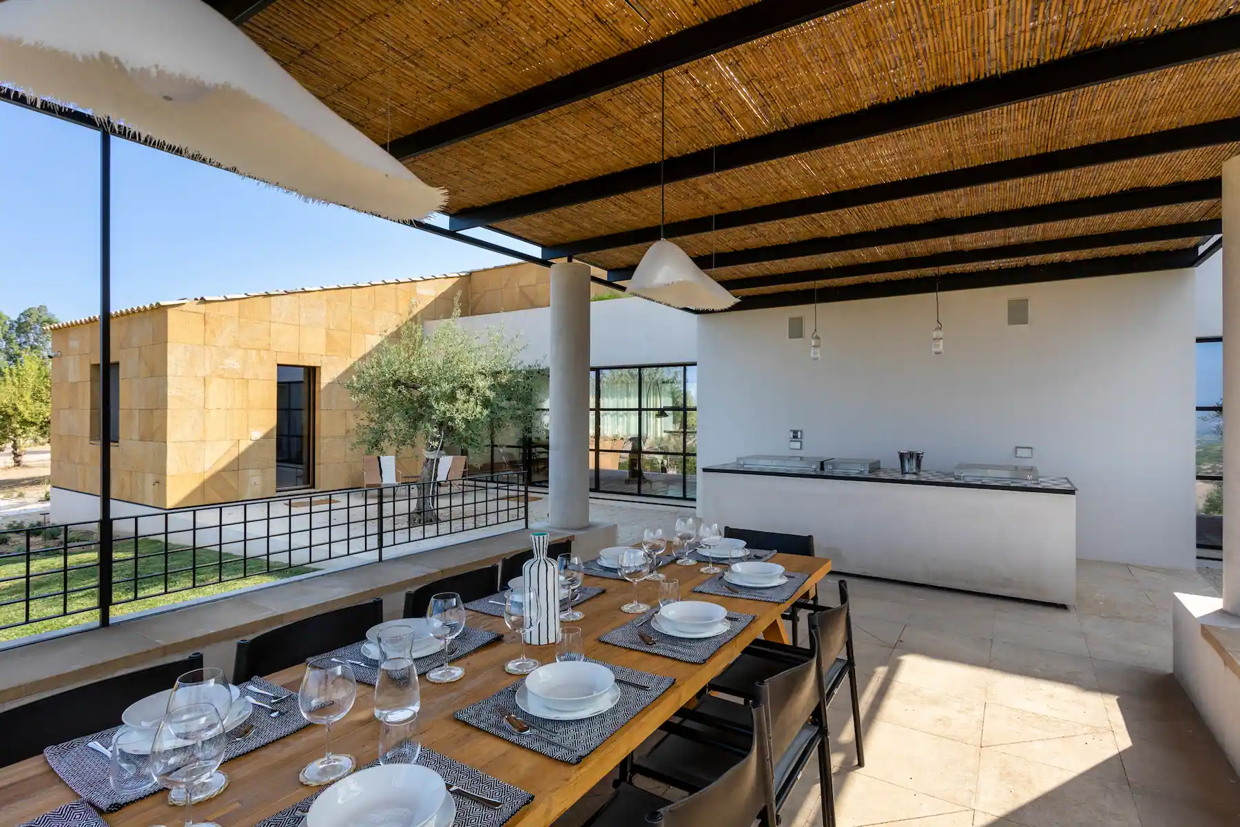 An outdoor dining area features a wooden table set for a meal, covered by a bamboo-roofed pergola. The table is set with plates, glasses, and cutlery. Behind the dining area is a modern building with large windows and beige stone walls. The sky is clear and blue.
