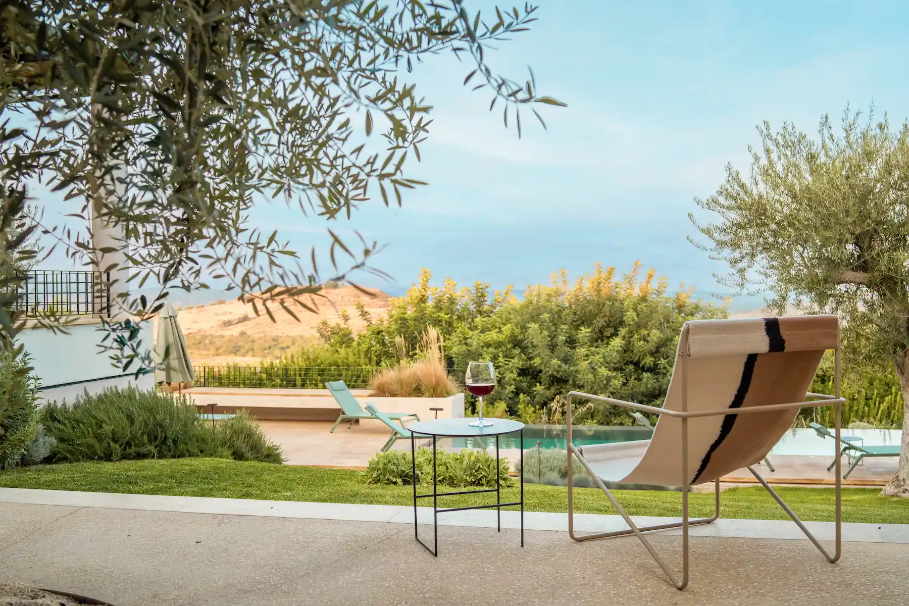 A serene outdoor setting with a lounge chair facing a panoramic view. A small table next to the chair holds a glass of red wine. In the background, a pool and lush vegetation are visible, with hills and trees under a calm blue sky.