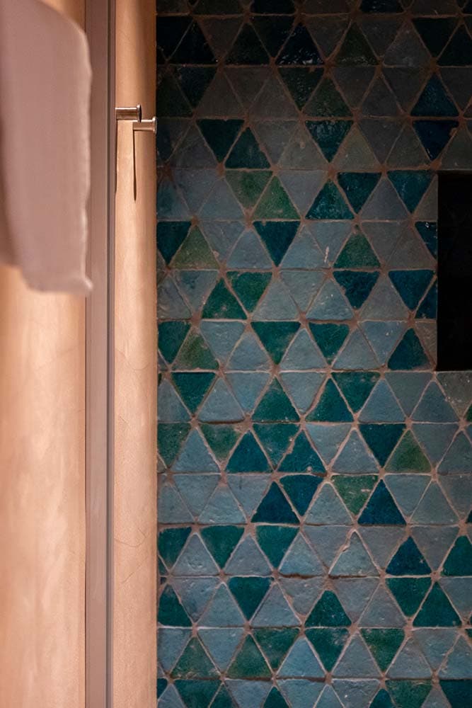 Un mur avec des carreaux triangulaires bleus et verts disposés selon un motif géométrique. À gauche, on peut voir une partie d'une poignée en métal et un mur de couleur claire. L'image montre un design moderne et élégant.