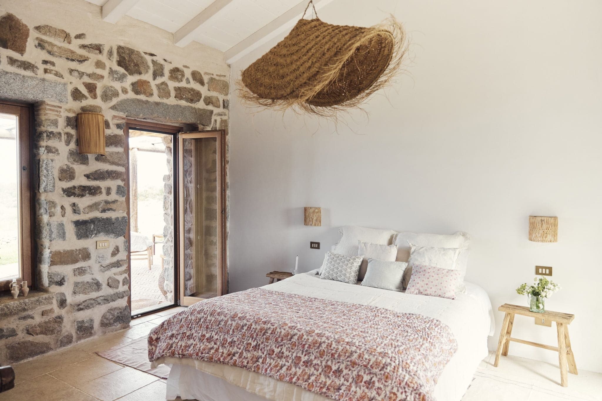 A cozy bedroom with a rustic design featuring a stone accent wall, a large bed with patterned bedding, wooden nightstands, and woven light fixtures. An open wooden door leads to an outdoor patio. A unique, large woven decorative piece hangs from the ceiling, adding character to the space.