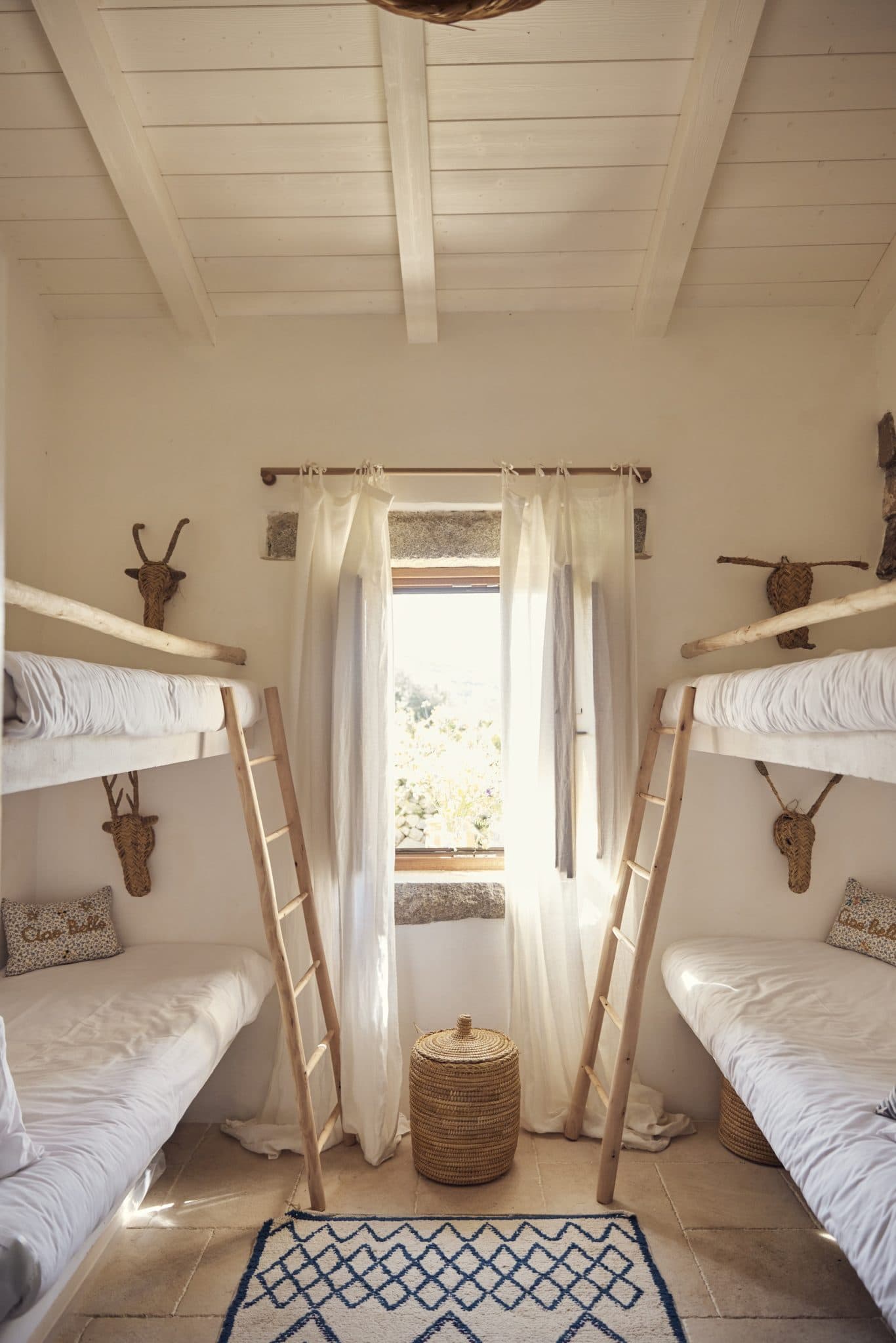 A cozy bedroom with two wooden bunk beds, each adorned with white bedding and cushions. A large window with sheer curtains allows natural light to stream in. A woven basket sits in the center of the room, and decorative animal heads are mounted on the white walls.
