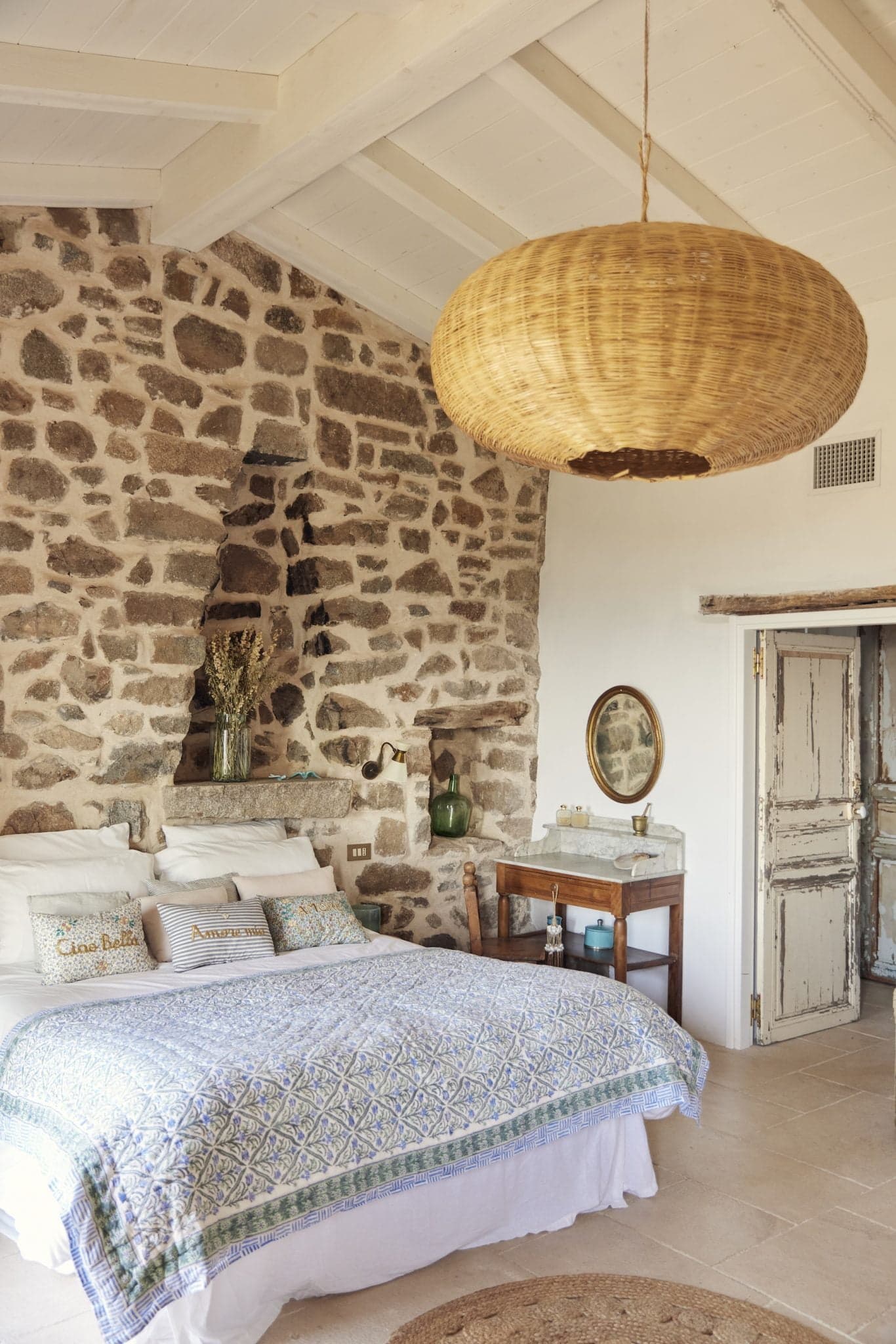 A cozy bedroom featuring an accent stone wall, a vaulted white ceiling, and a large wicker pendant light. The bed, with patterned bedding, is set against the wall next to a rustic nightstand holding a mirror and decorative items. An old wooden door is partially open on the right.